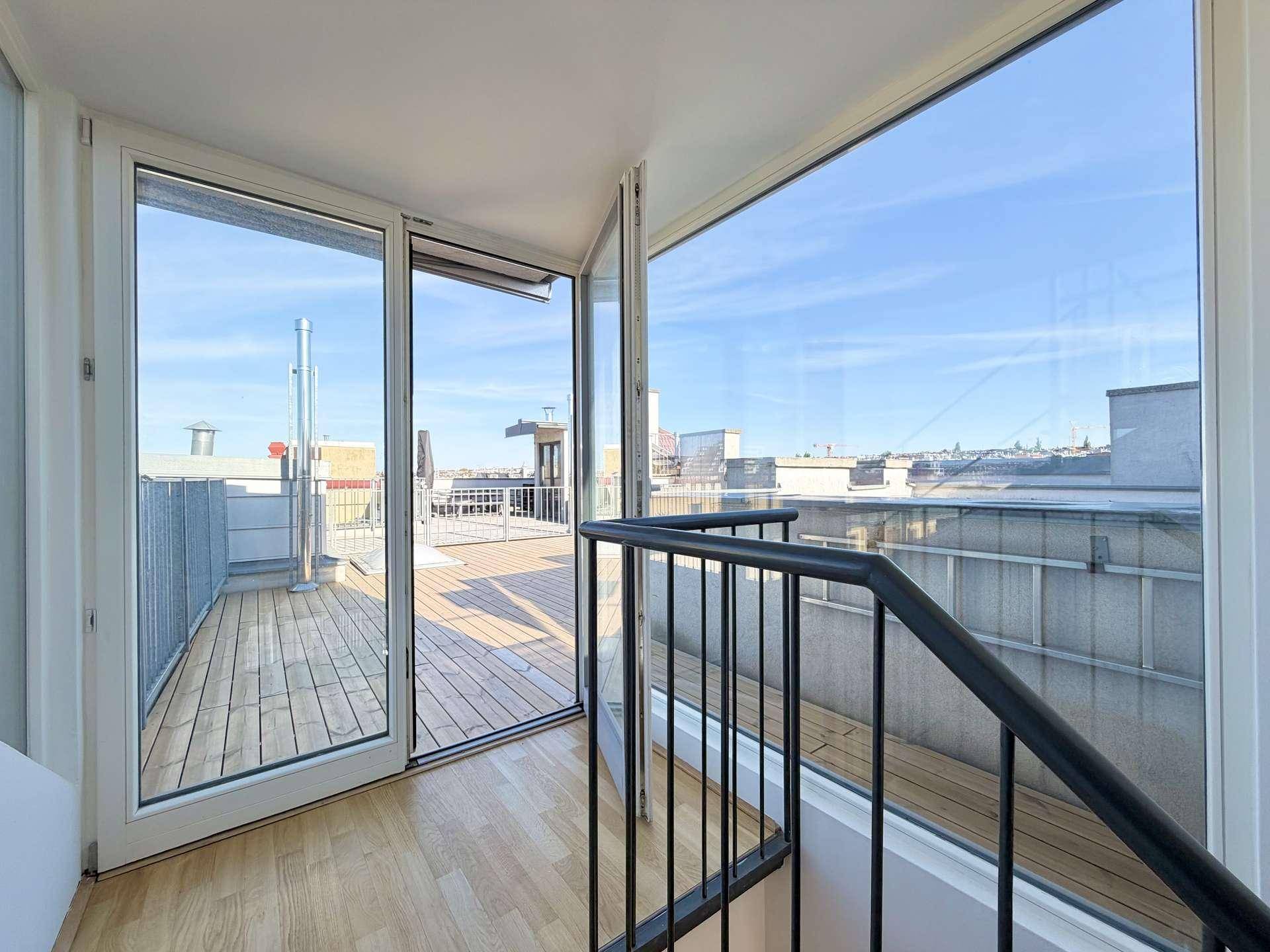 Innenansicht mit Blick durch große Glasfront auf die Dachterrasse und die Stadt.