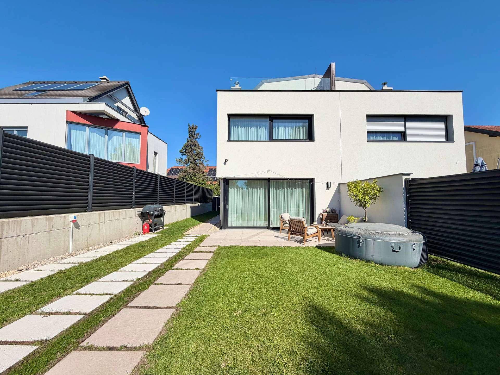 Rückansicht des Hauses mit sonnigem Garten, Terrasse und Whirlpool, umgeben von einem Zaun.
