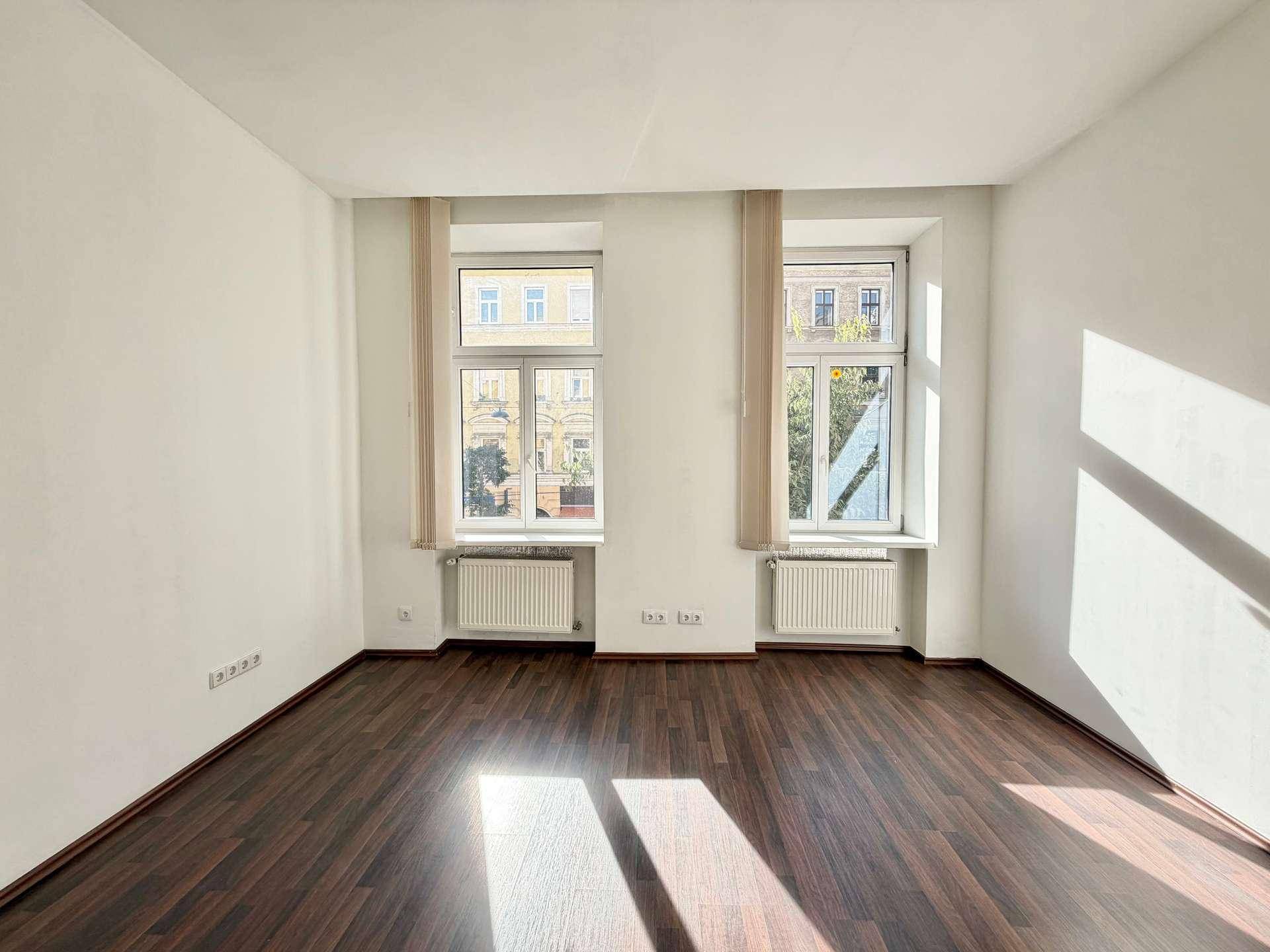 Leeres Wohnzimmer mit Laminatboden, weißen Wänden und zwei großen Fenstern mit viel Licht.