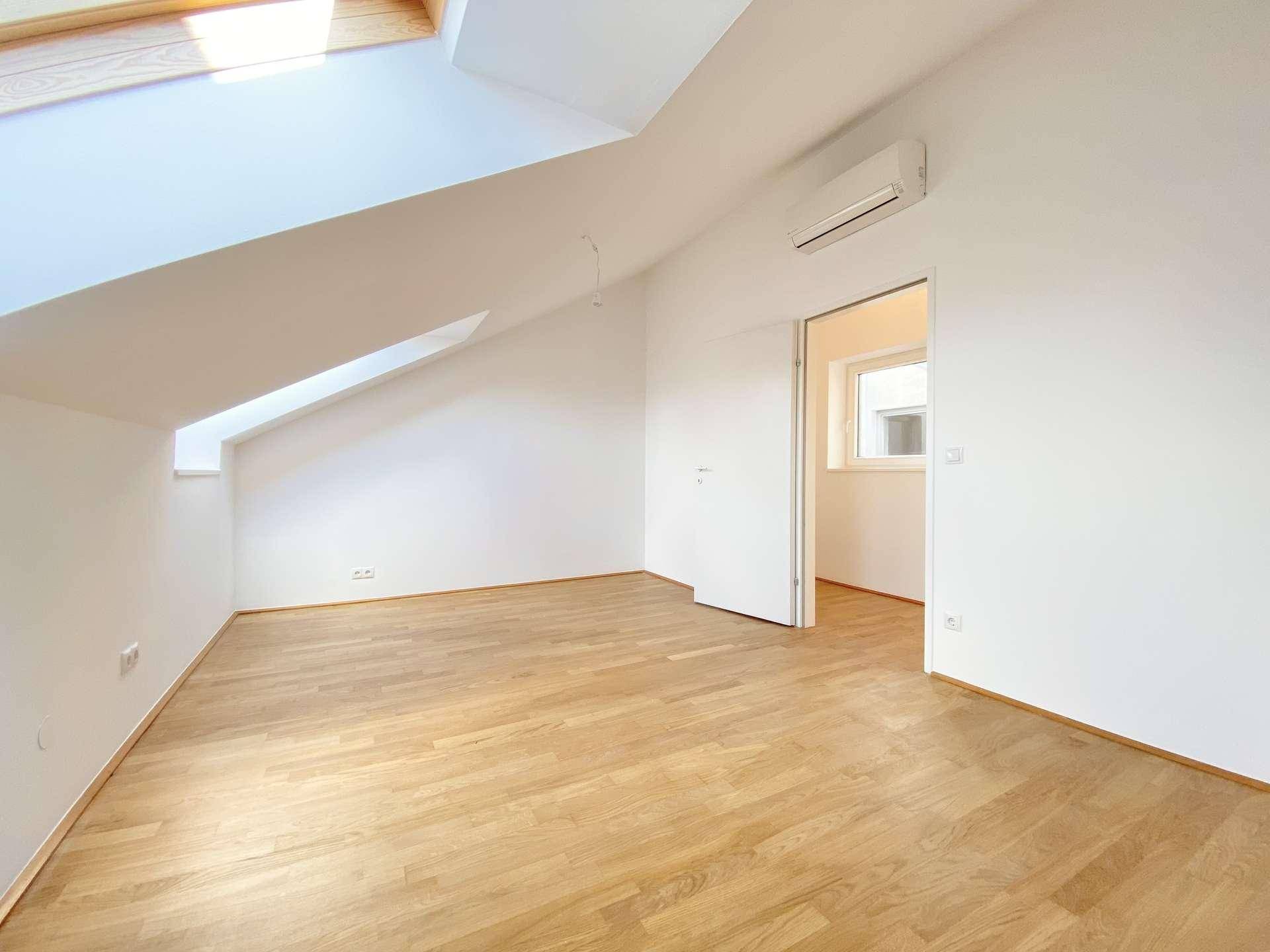 Helles Zimmer mit Holzfußboden, schrägen Wänden, Dachfenster und Klimaanlage.