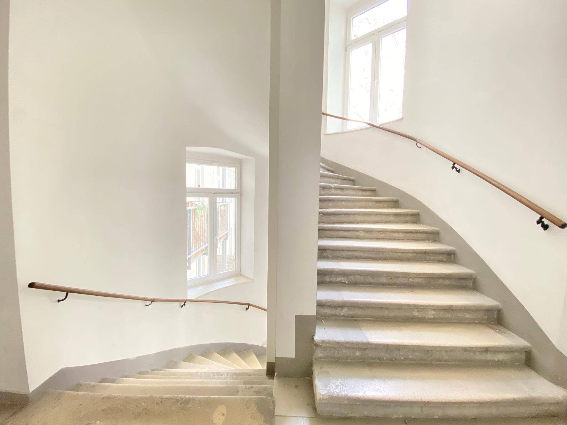 Helles Treppenhaus mit geschwungener Steintreppe und Holzhandläufen, gut beleuchtet durch große Fenster.