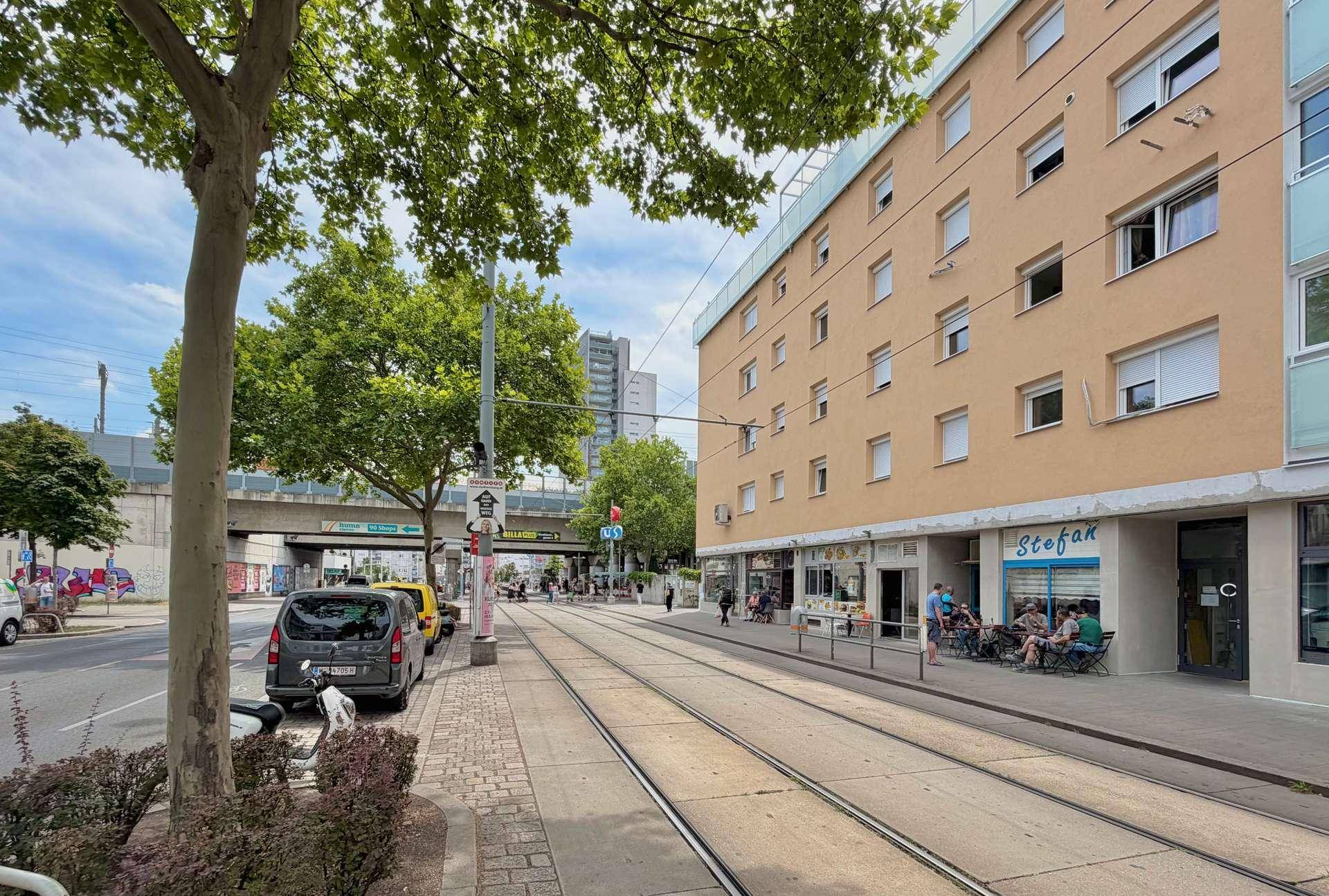 Straßenansicht eines Wohngebäudes mit Geschäften im Erdgeschoss und Straßenbahnschienen davor.