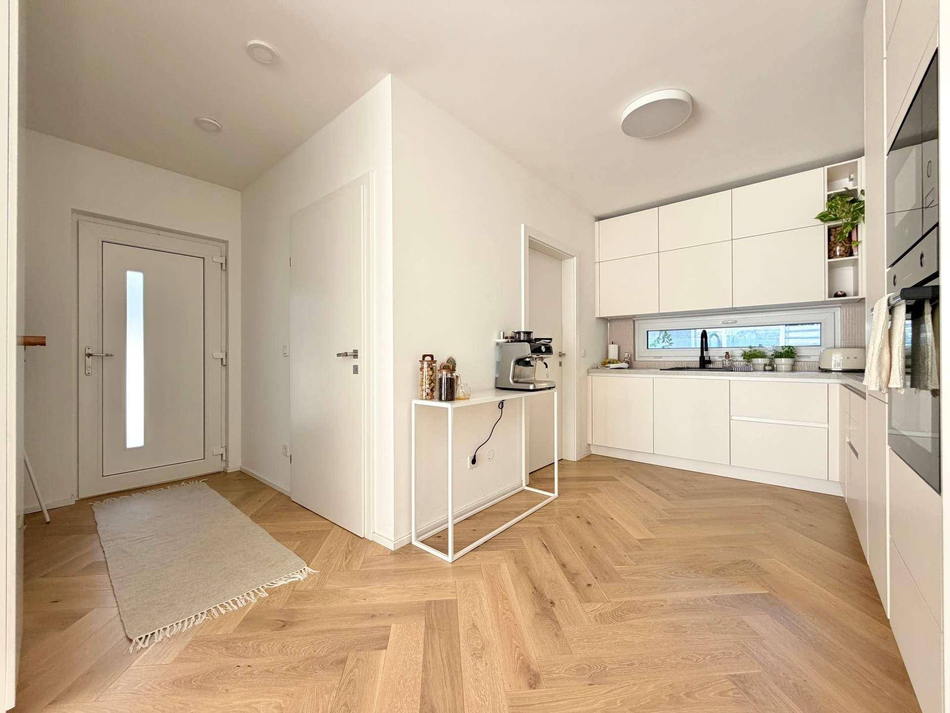 Moderne Küche mit weißen Schränken, integrierten Geräten und Holzboden im Eingangsbereich.