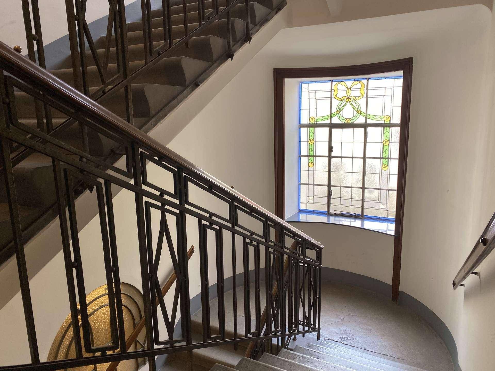 Historisches Treppenhaus mit kunstvollem Geländer und farbigem Bleiglasfenster.