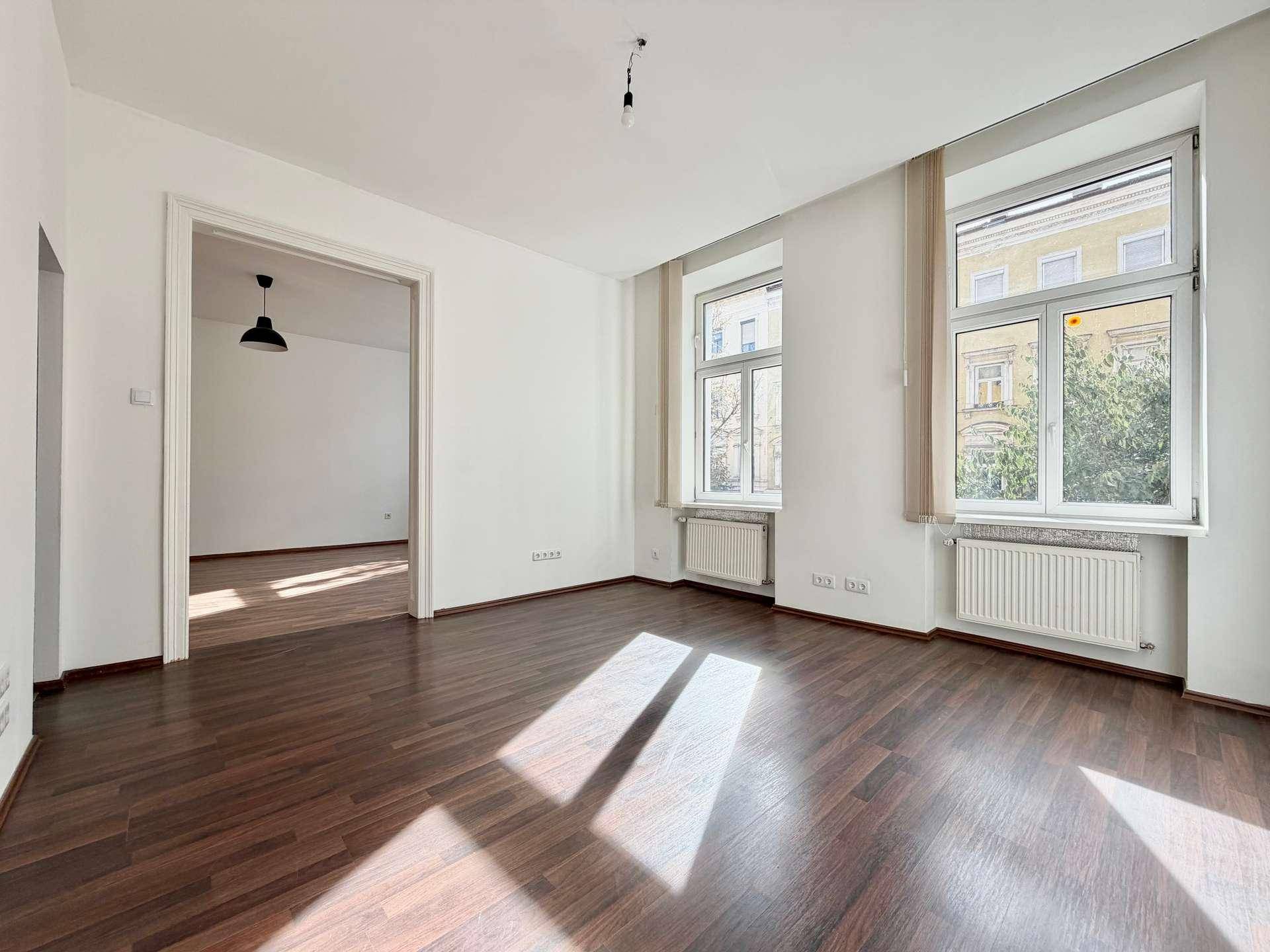 Helles Wohnzimmer mit Laminatboden und Blick auf die Straße, verbunden mit einem weiteren Raum.
