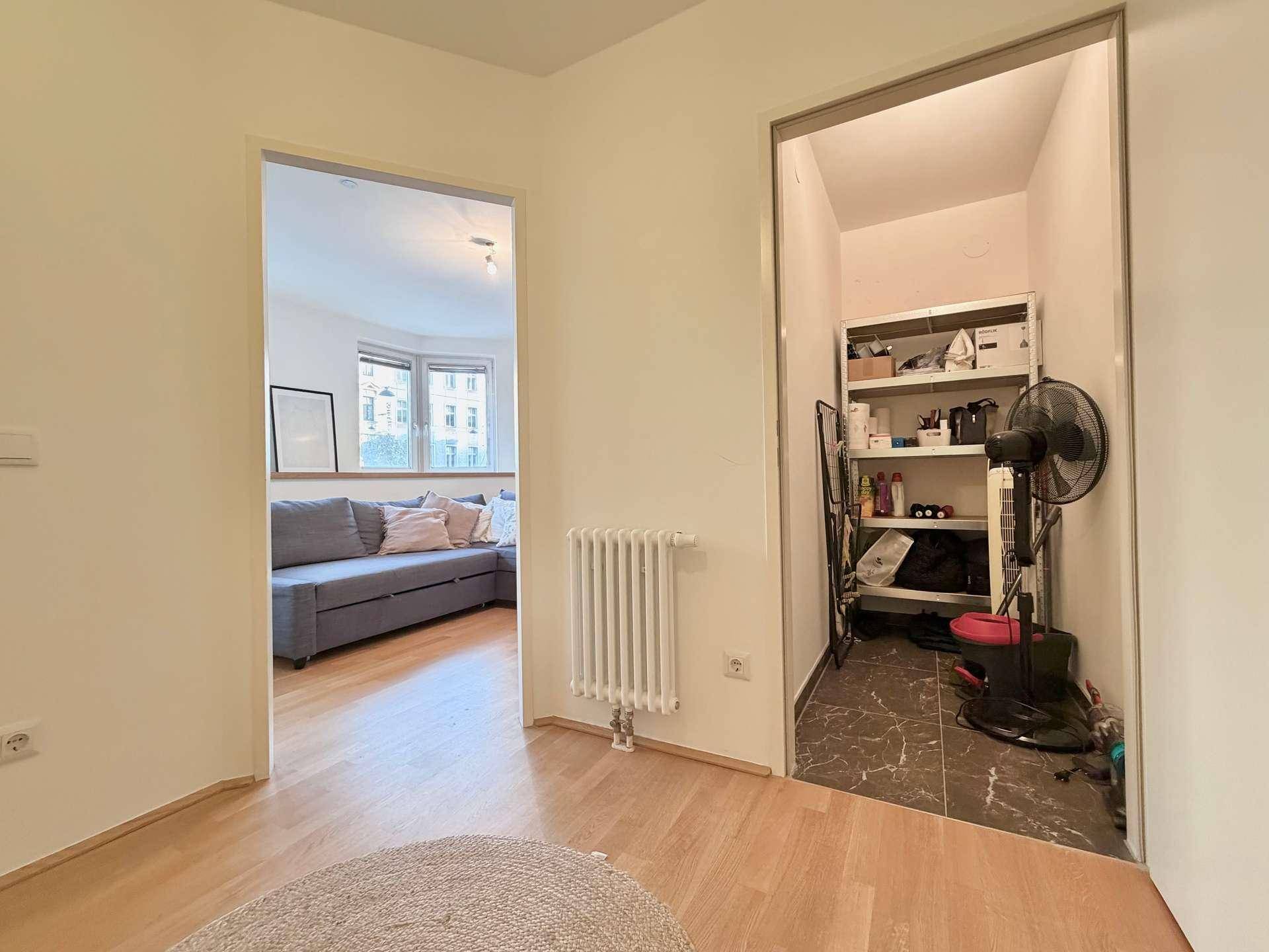 Blick von einem Flur in ein helles Wohnzimmer und einen praktischen Abstellraum mit Regalen.