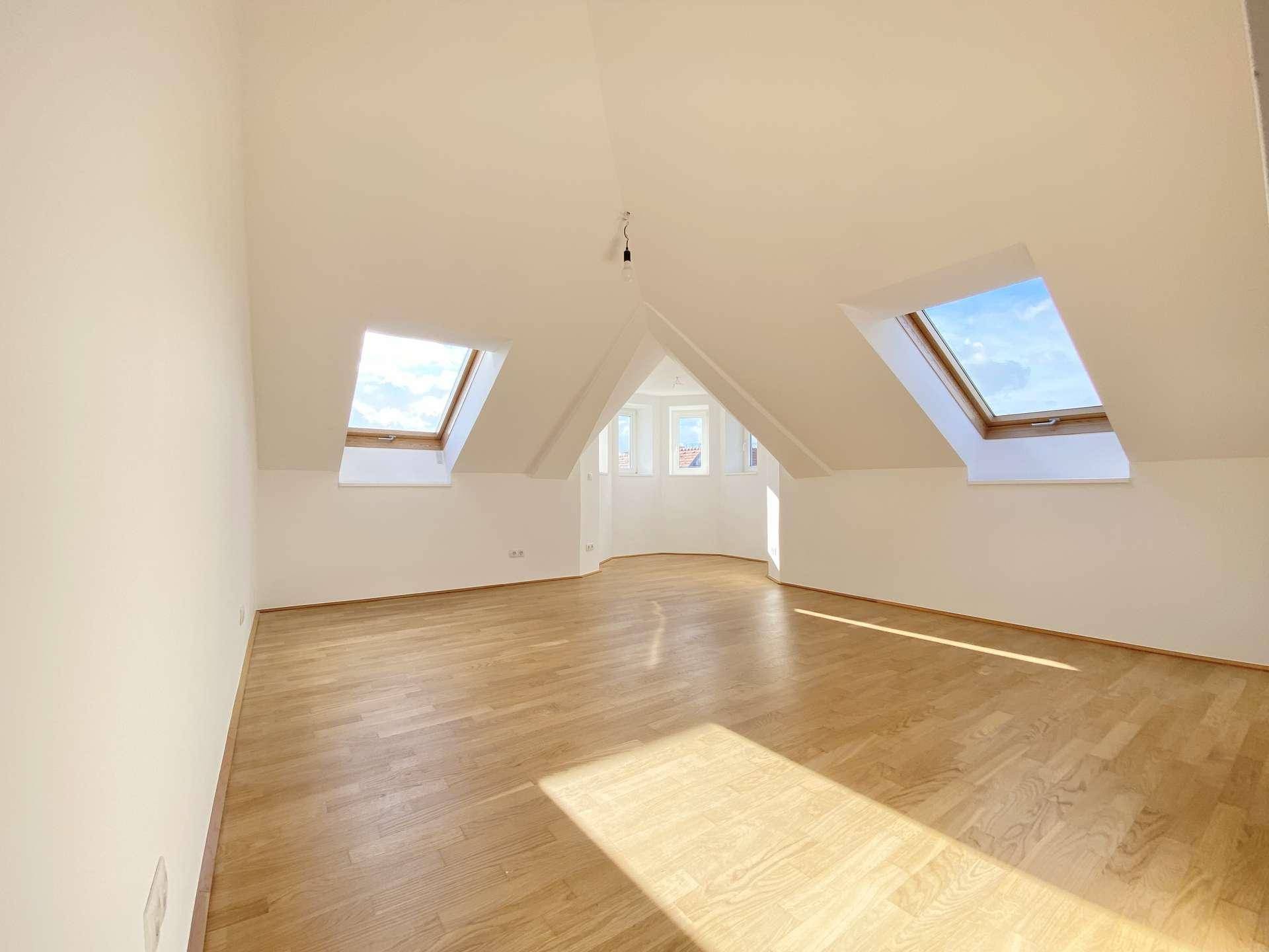 Großes, helles Zimmer mit Holzfußboden, schrägen Wänden und mehreren Dachfenstern.