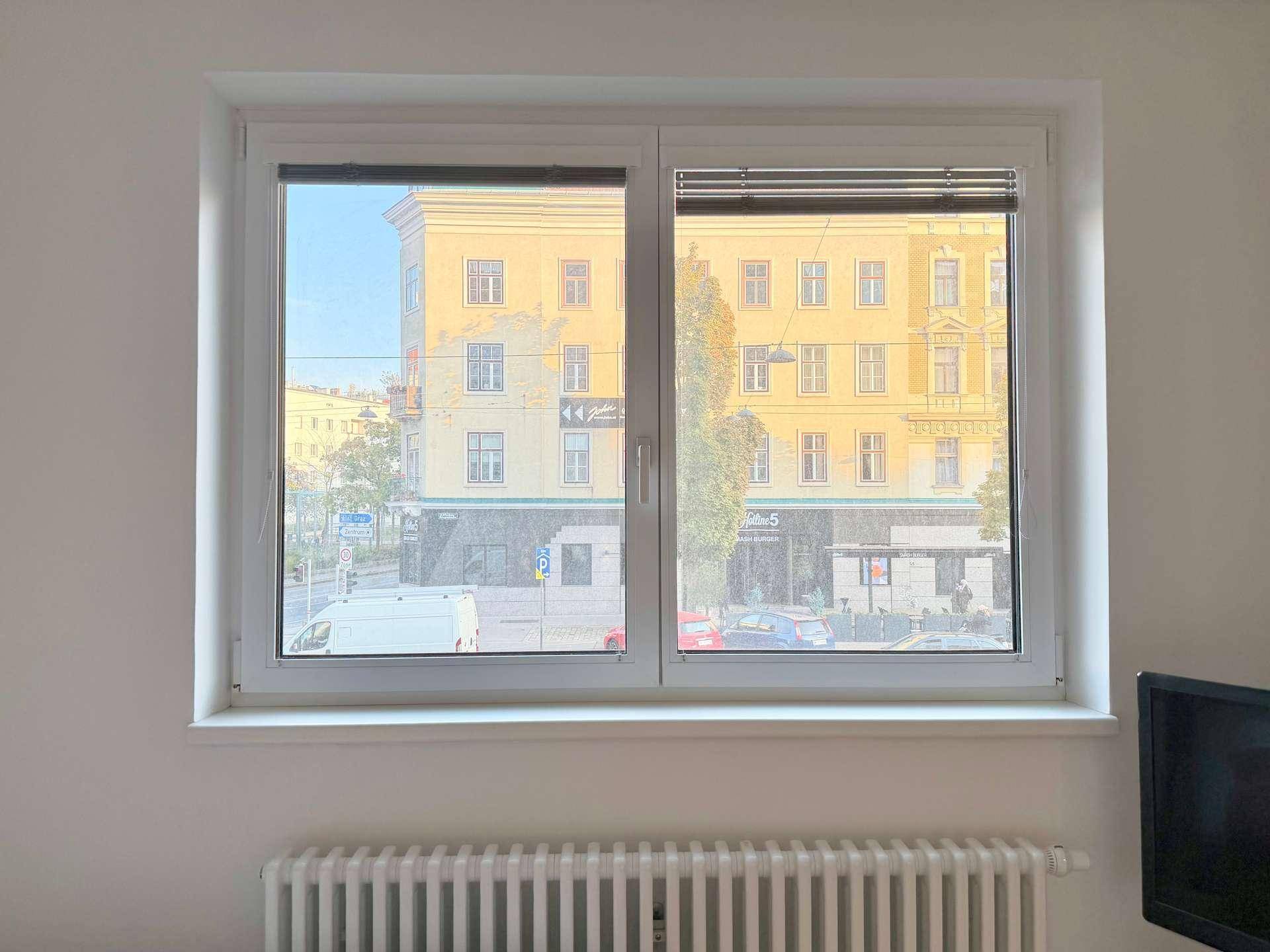 Fenster mit Blick auf eine belebte Stadtstraße und gegenüberliegende Gebäude bei Tageslicht.