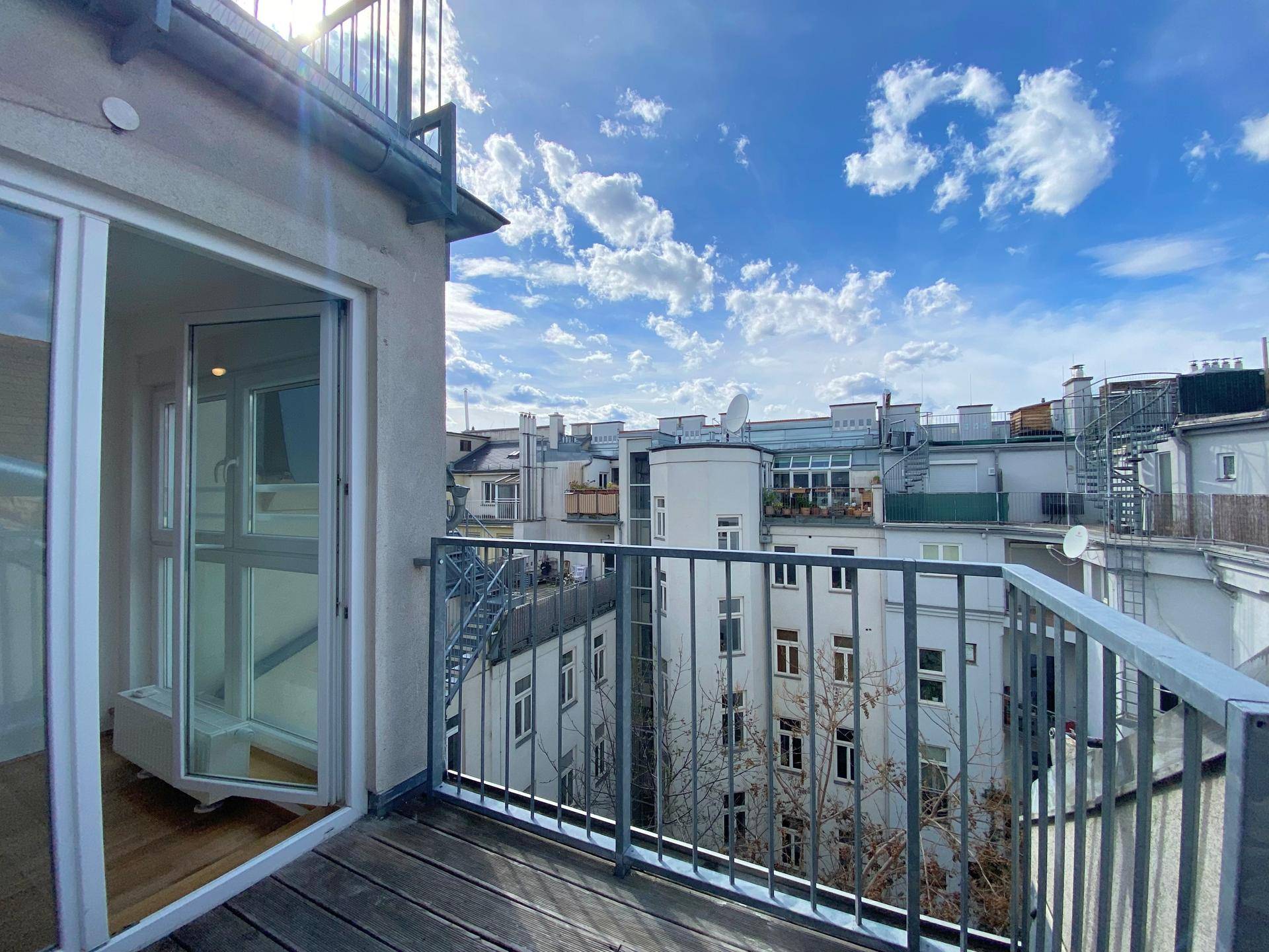 Sonniger Balkon mit Holzboden und Metallgeländer, bietet einen weiten Blick über die Dächer der Stadt.