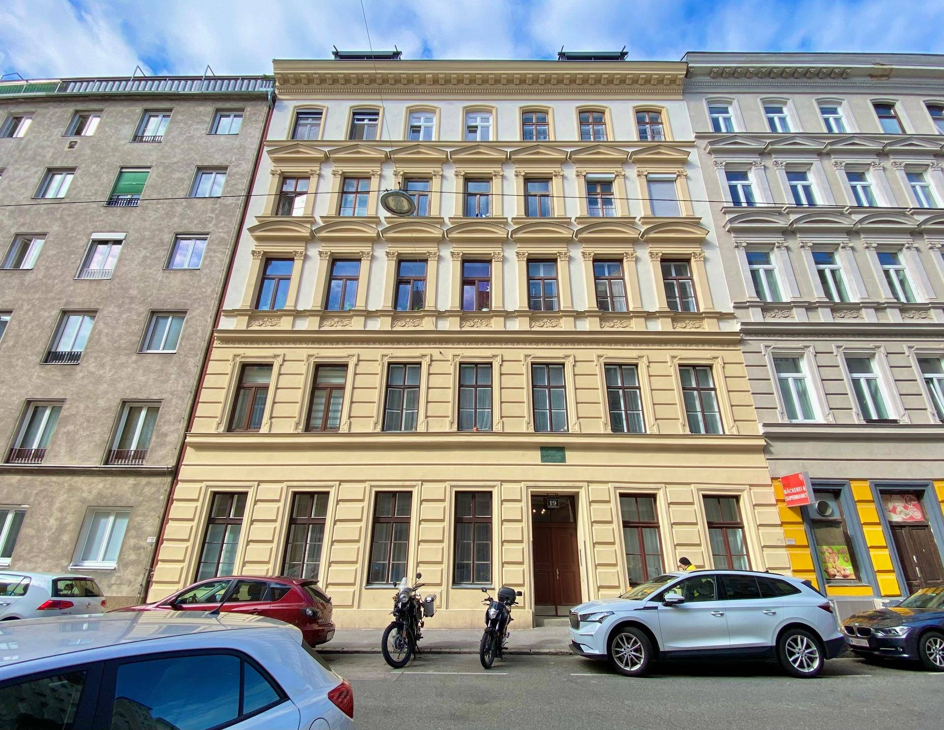 Klassische Fassade eines Mehrfamilienhauses mit vielen Fenstern und parkenden Autos auf der Straße davor.