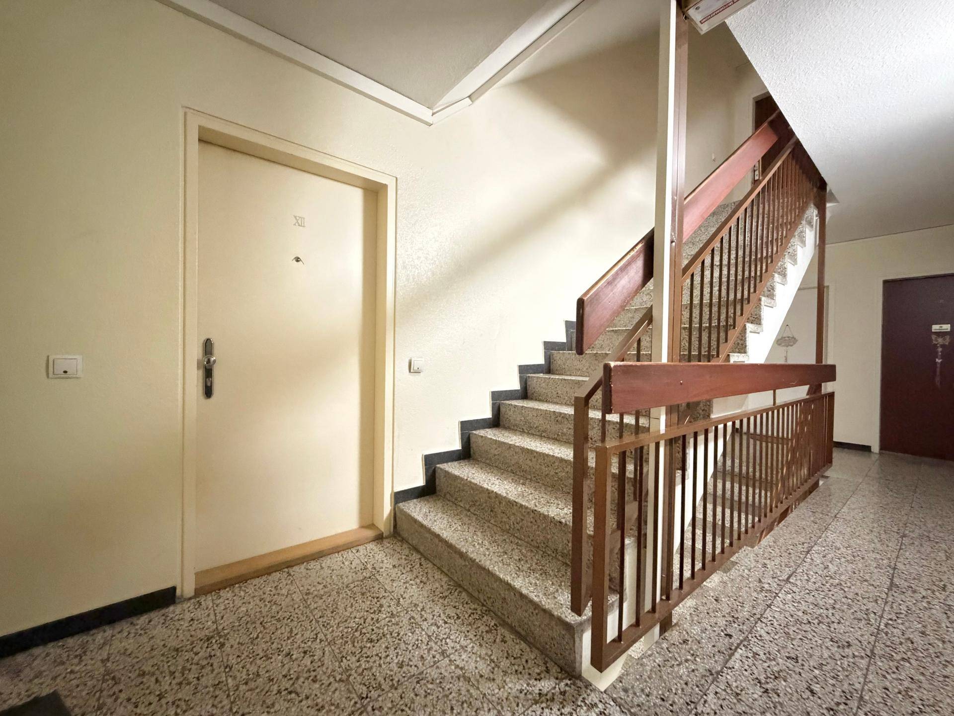 Treppenhaus mit Terrazzoboden und Holzgeländer, das zu den oberen Etagen führt.