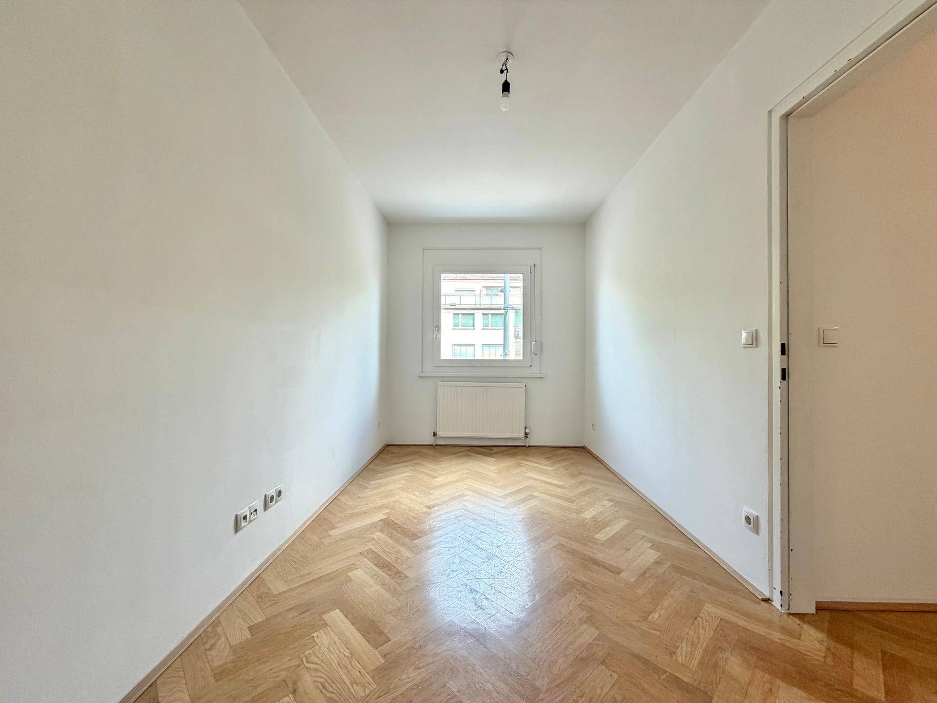 Geräumiges Zimmer mit Fischgrätparkett und einem Fenster, das viel Tageslicht hereinlässt.