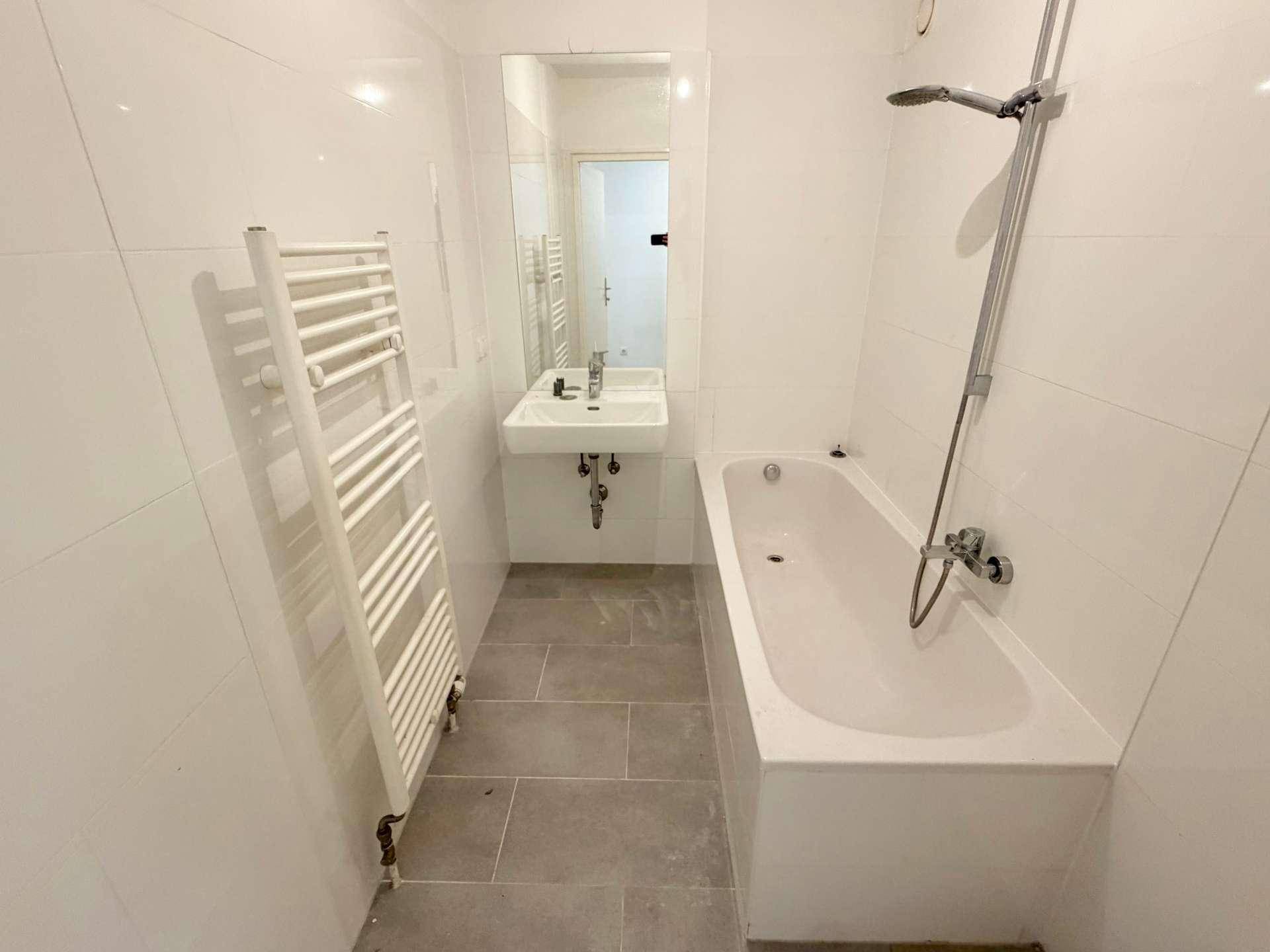 Modernes Badezimmer mit weißen Wandfliesen, grauen Bodenfliesen, Badewanne und Handtuchheizkörper.