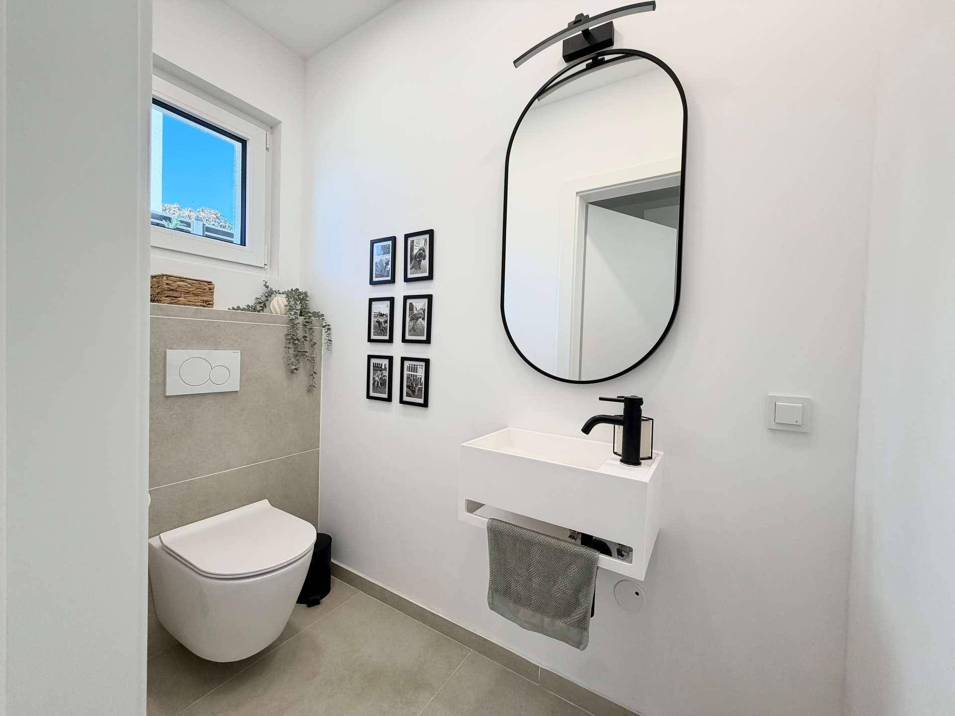 Modernes Gäste-WC mit wandhängender Toilette, kleinem Waschbecken und ovalem Spiegel.