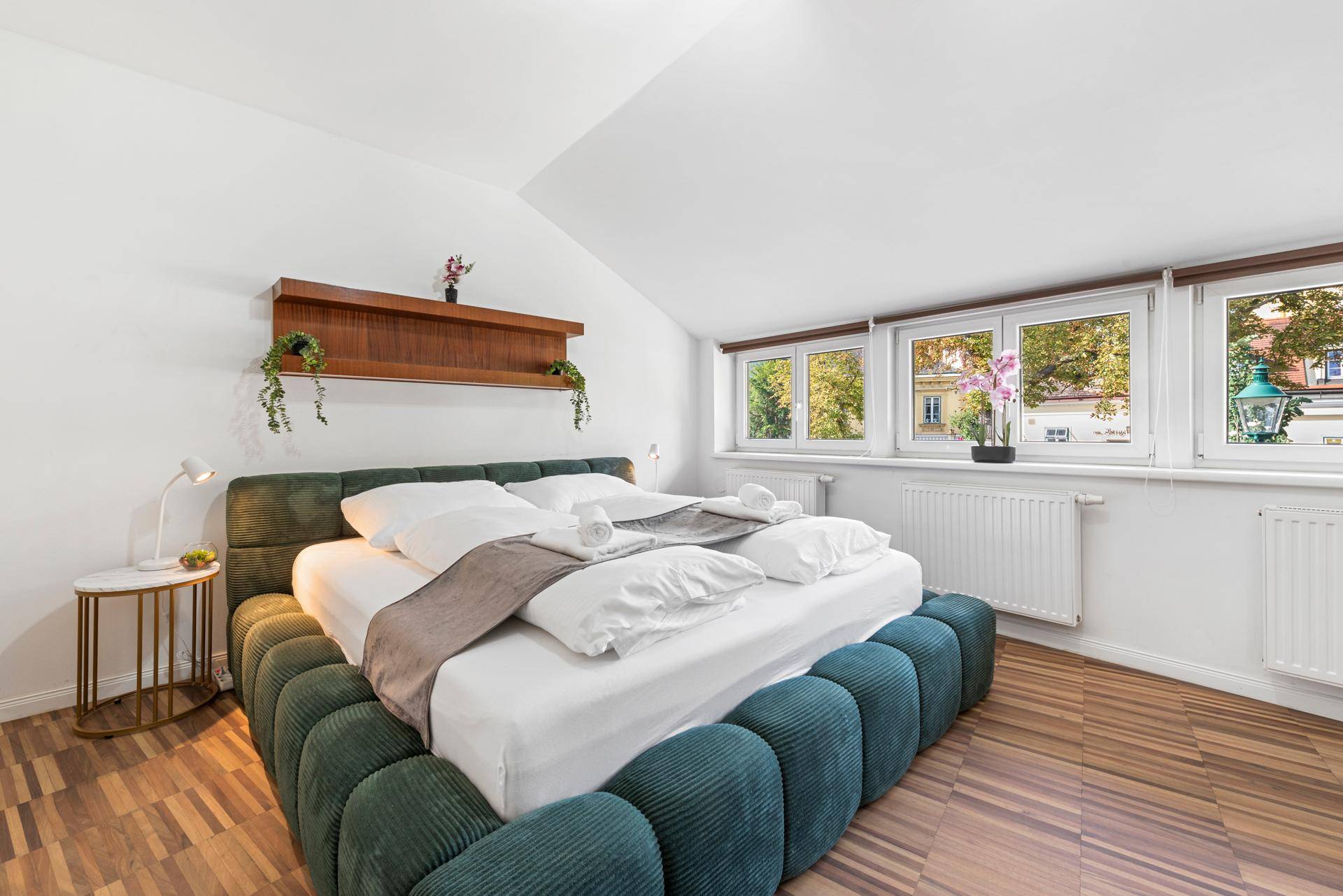 Helles Schlafzimmer im Dachgeschoss mit grünem Bett und Fenstern mit Blick nach draußen.
