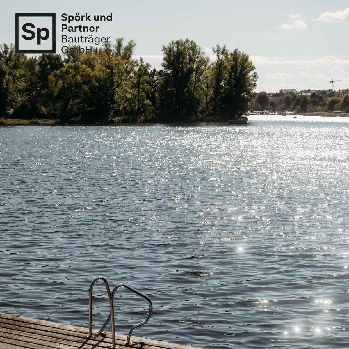 Weitläufiger Blick auf einen sonnigen See mit bewaldetem Ufer und einem kleinen Badesteg.