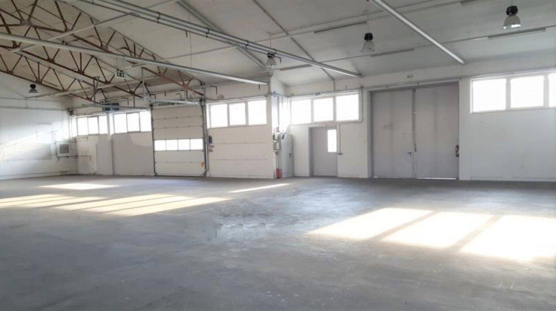 Geräumige, leere Lagerhalle mit Betonboden, hohen Decken und viel Tageslicht durch große Fenster.