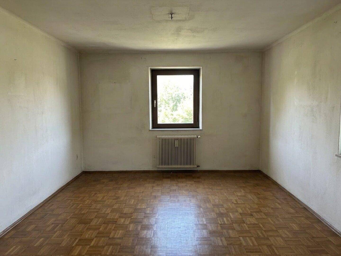 Leerer Raum mit hellem Parkettboden, einem Fenster und einem Heizkörper darunter.