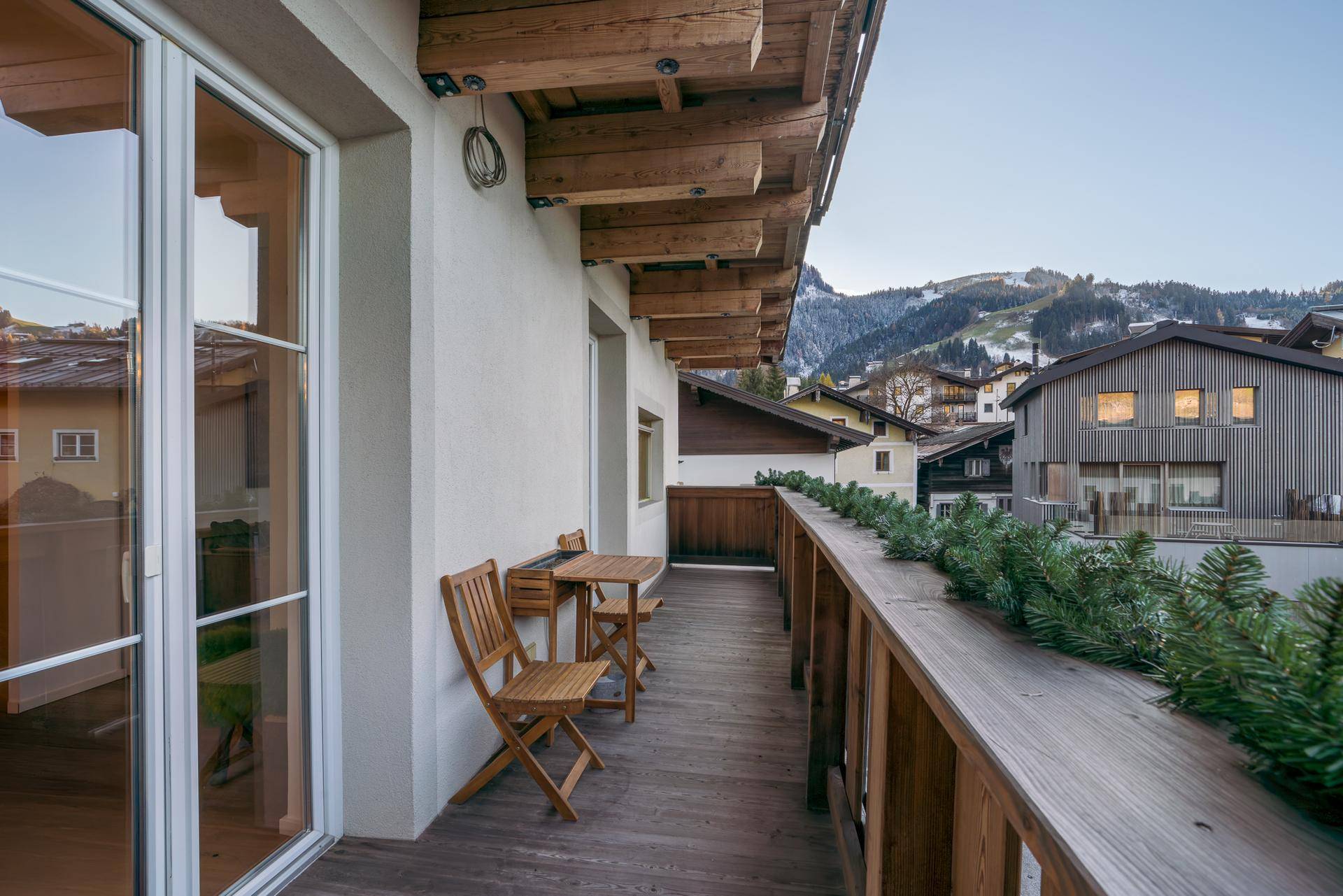 Langer Holzbalkon mit Sitzgelegenheiten und Blick auf die umliegende Berglandschaft und Häuser.