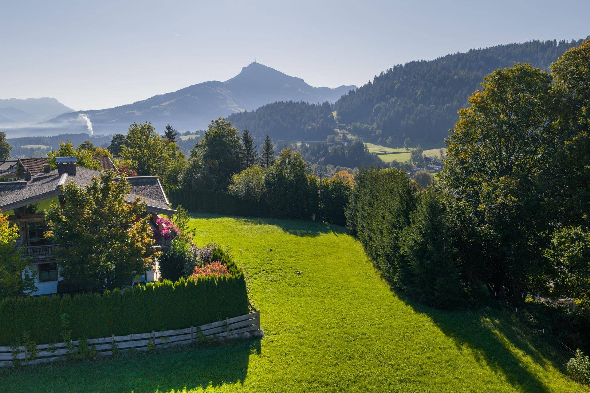 Seitenansicht eines Hauses mit gepflegtem Gartenbereich, umgeben von grünen Hügeln und Bäumen.