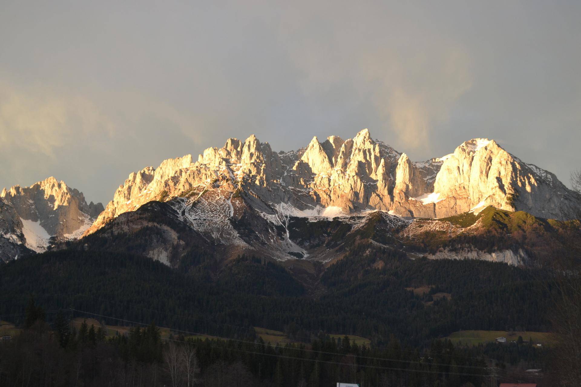 Majestätische, schneebedeckte Berge im warmen Licht der untergehenden Sonne, umgeben von dunklen Wäldern.