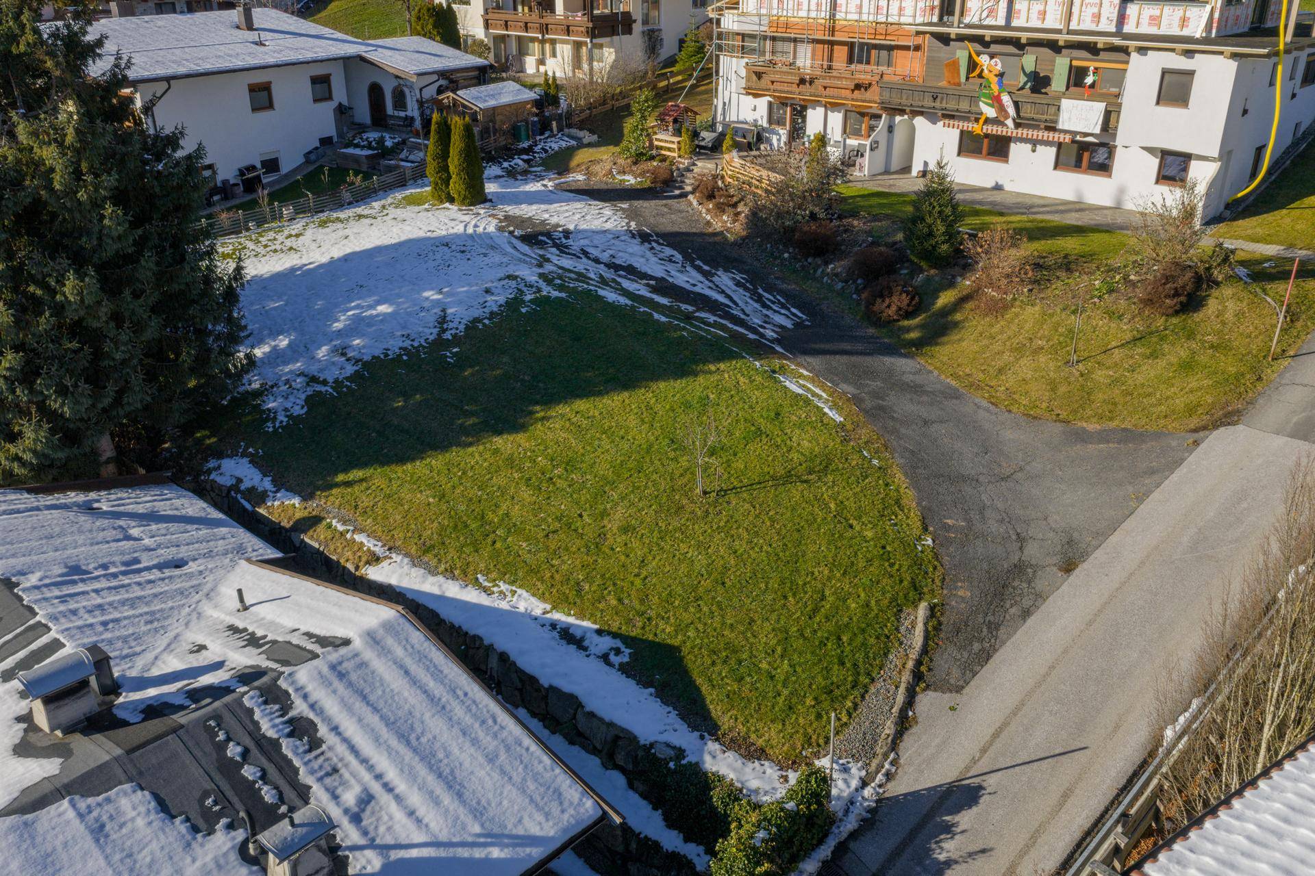 Grundstück mit grünen Grasflächen und Schneeresten, umgeben von Wohnhäusern und einer Zufahrt.