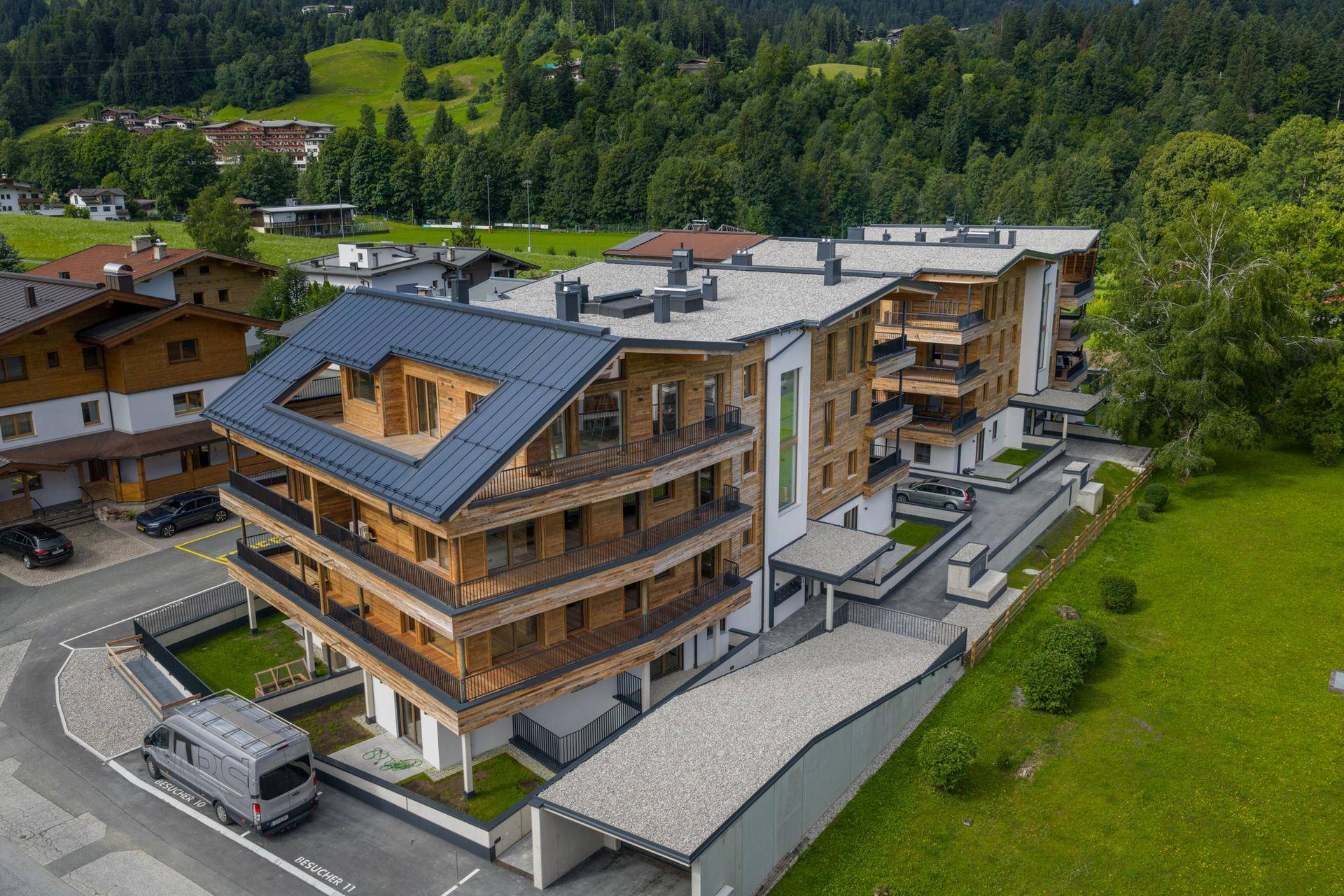 Luftaufnahme eines modernen Apartmenthauses mit Holzfassade und Balkonen, umgeben von grünen Hügeln.