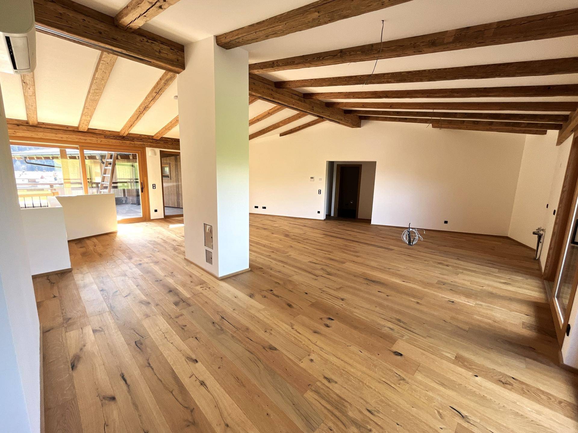 Heller Wohnbereich mit sichtbaren Holzbalken, Holzboden und Zugang zu weiteren Räumen.
