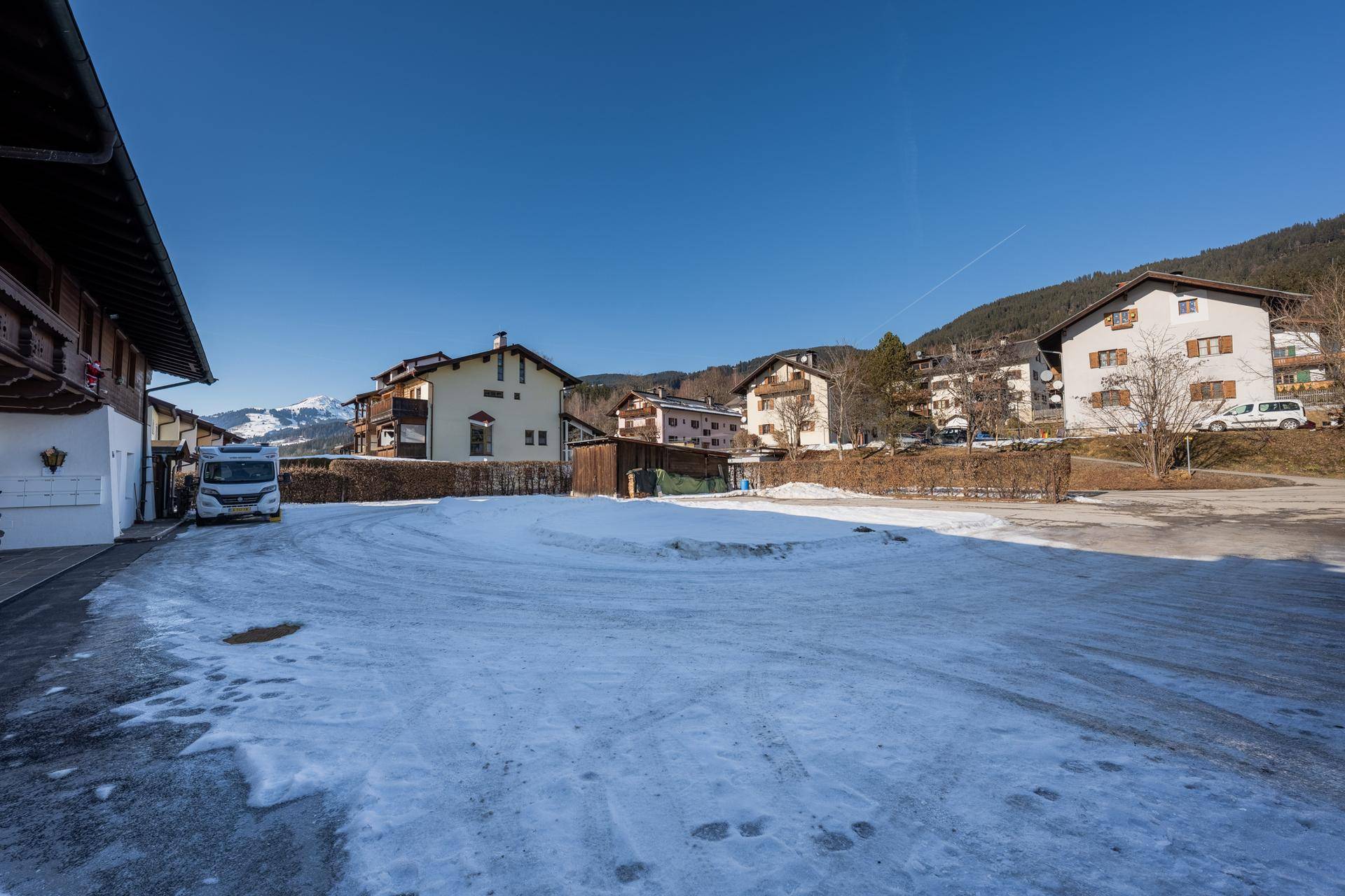 Weitläufiger, verschneiter Parkplatz vor einer Reihe von Häusern in einer bergigen Landschaft unter blauem Himmel.
