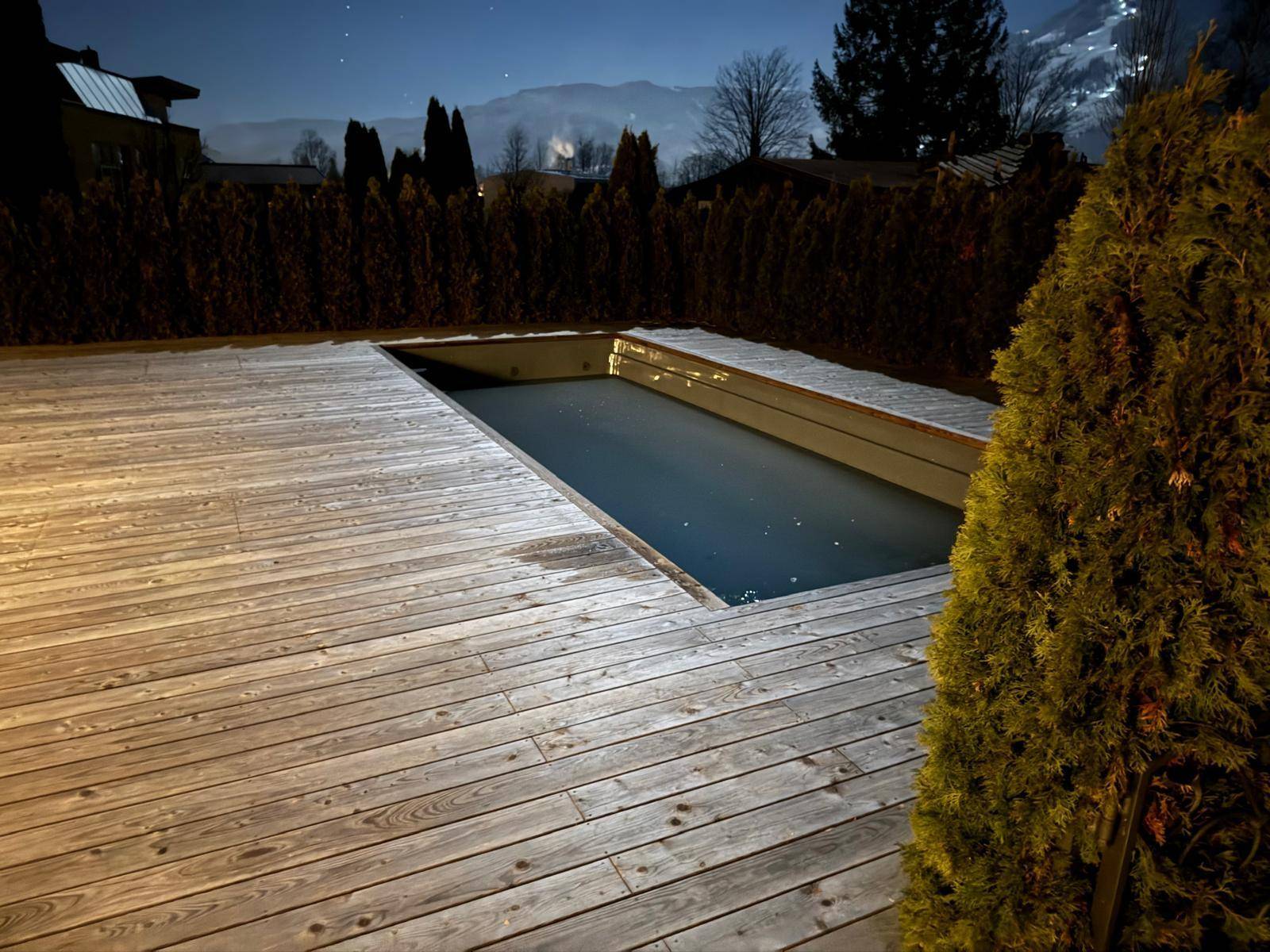 Außenpool mit Holzdeck bei Nacht, umgeben von Hecken und Bäumen.