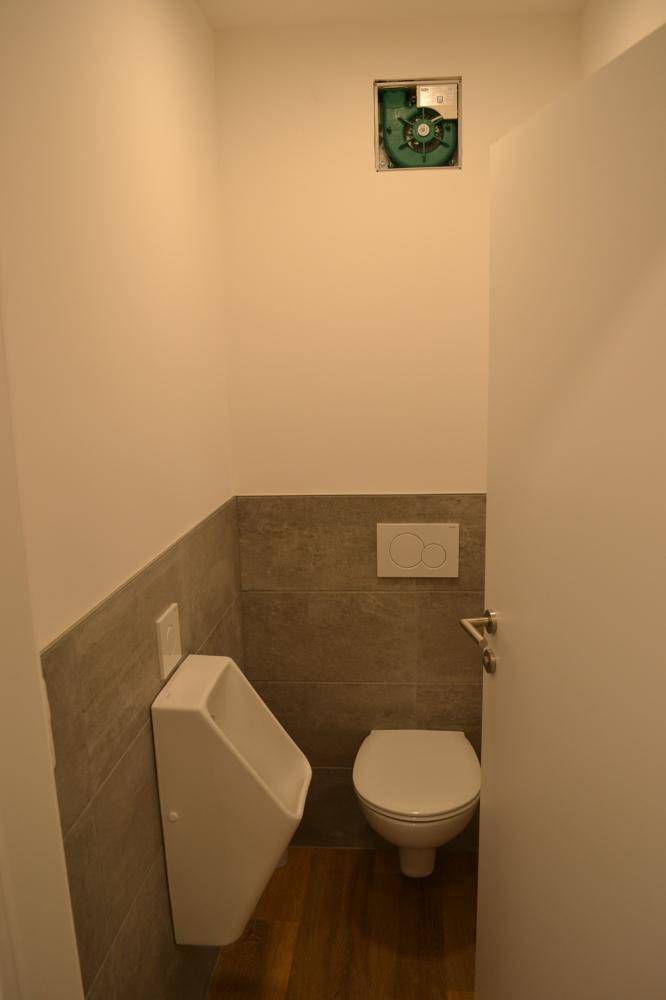 Kleiner Toilettenraum mit Urinal, WC, grauen Fliesen und einem Abluftventilator an der Decke.