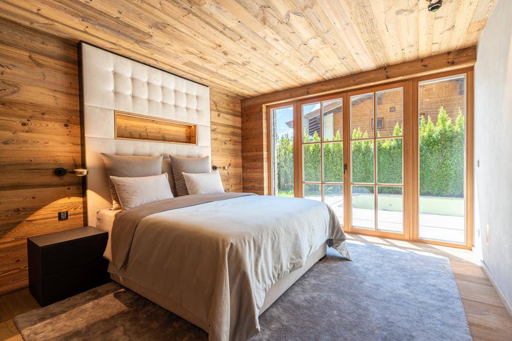 Schlafzimmer im Chalet-Stil mit Holzwänden, gepolstertem Kopfteil und großen Fenstern zum Garten.