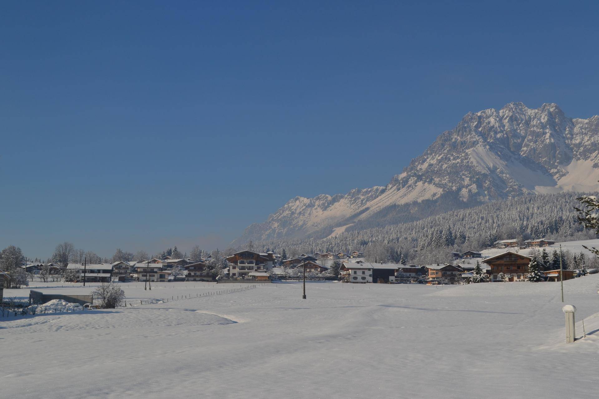 Verschneite Dorflandschaft in einem Tal, umgeben von weiten Feldern und majestätischen Bergen im Winter.