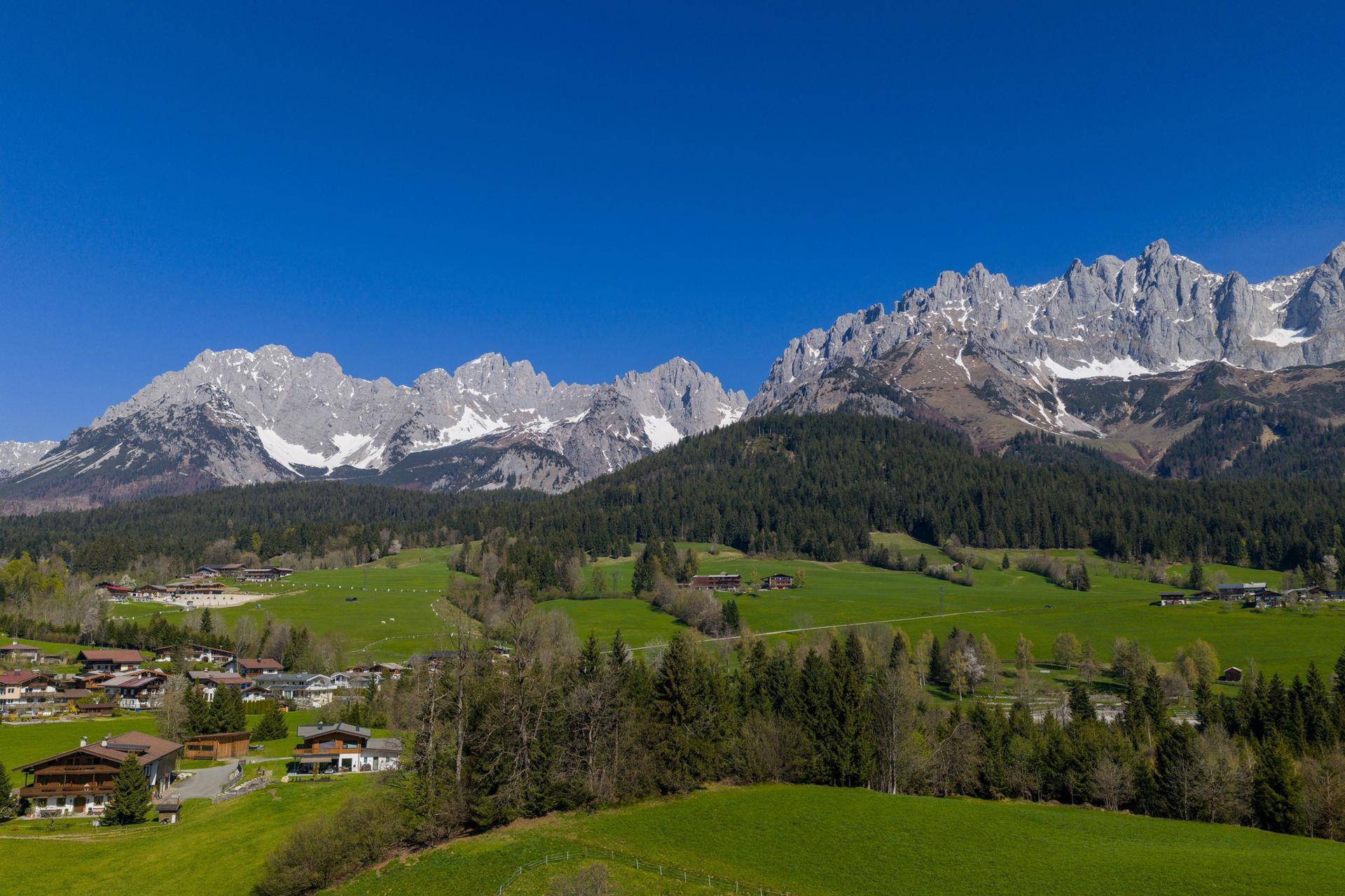 Weitläufige Landschaft mit grünen Wiesen, Wäldern und traditionellen Häusern vor imposanter Bergkulisse unter blauem Himmel.