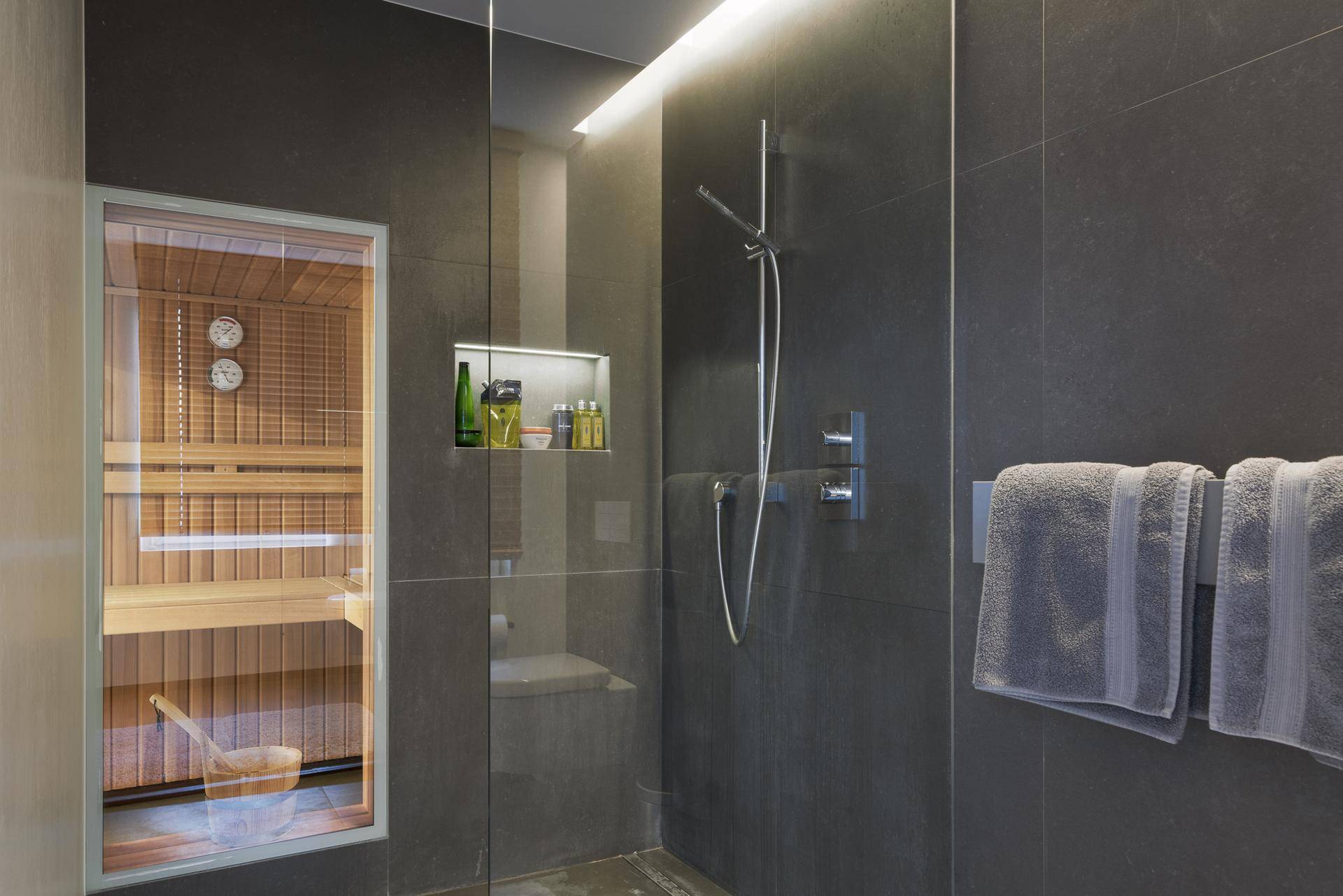Modernes Badezimmer mit begehbarer Dusche und Blick auf eine private Sauna, ausgestattet mit dunklen Fliesen.