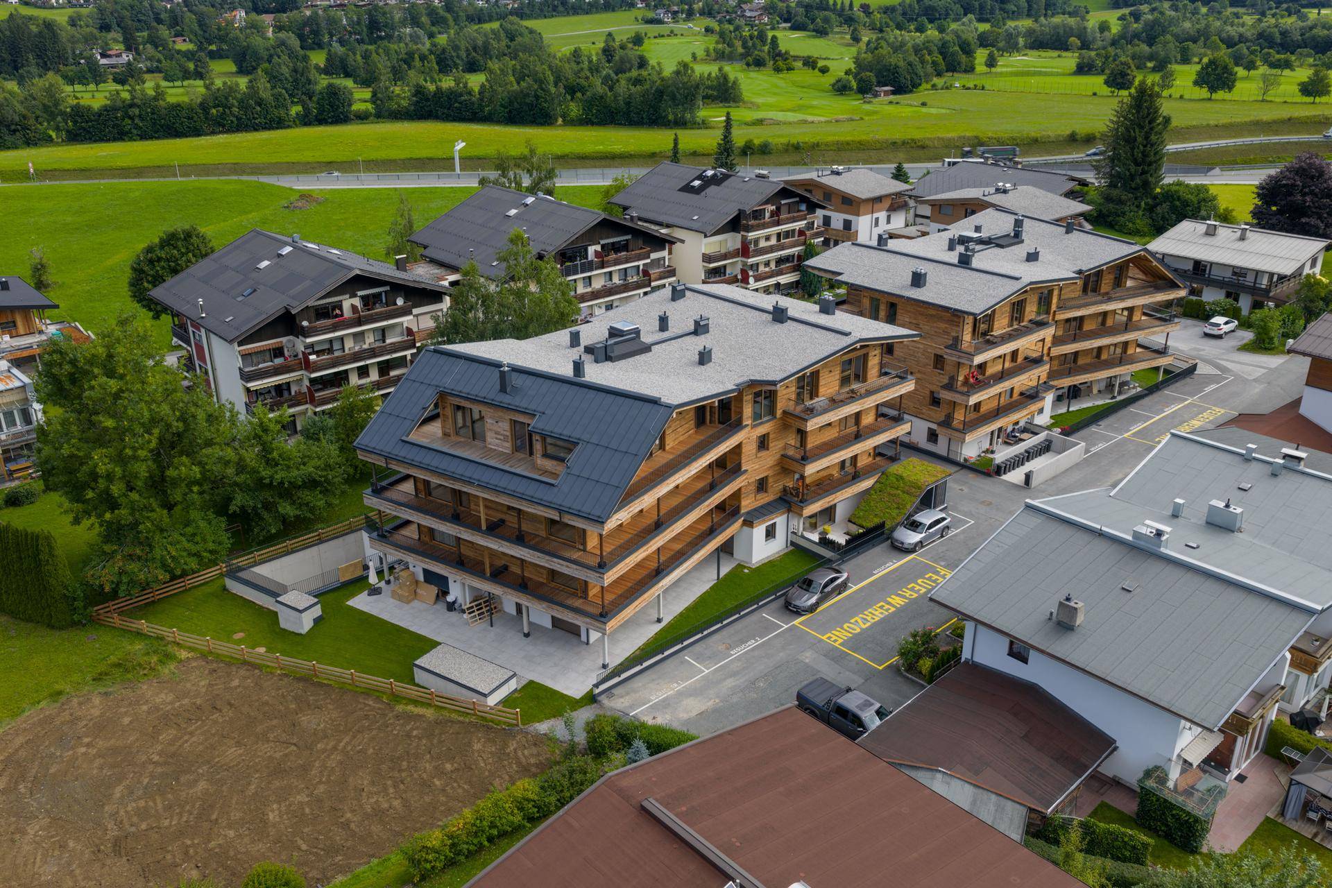 Luftaufnahme eines modernen Wohnkomplexes mit Holzfassaden und Balkonen, umgeben von grünen Wiesen und Bergen.