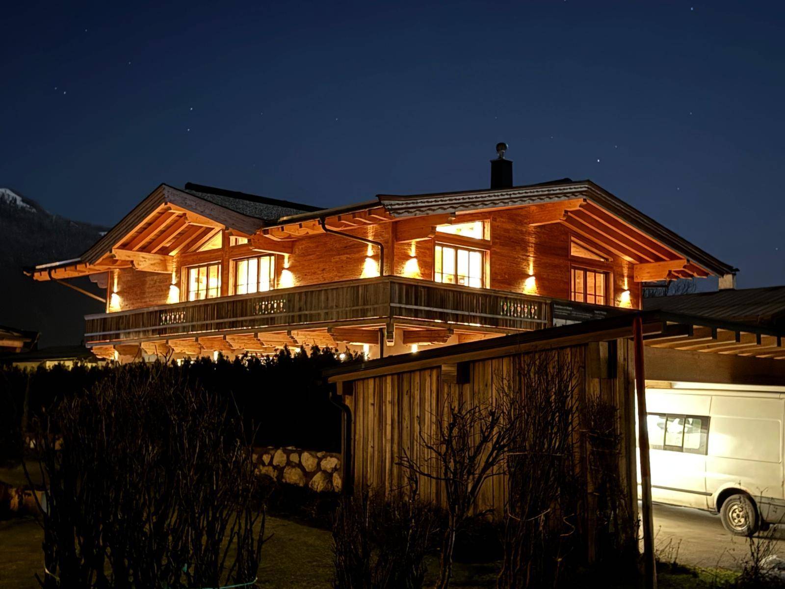 Beleuchtetes Holzchalet bei Nacht mit großem Balkon und Bergblick im Hintergrund.