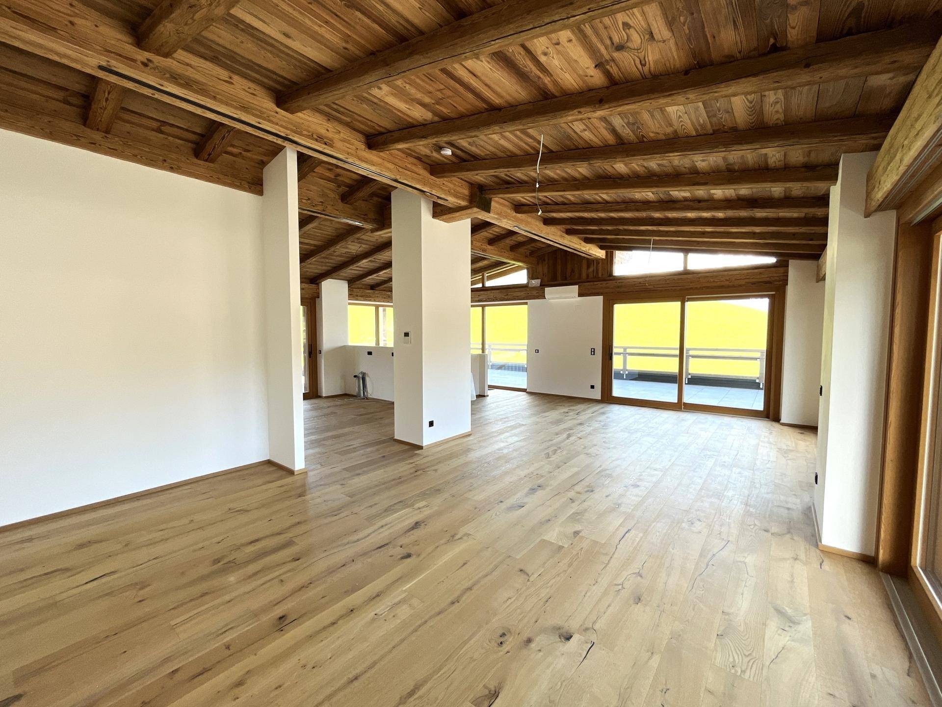 Offener Wohnbereich mit sichtbaren Holzbalken, Holzboden und großen Fenstern.