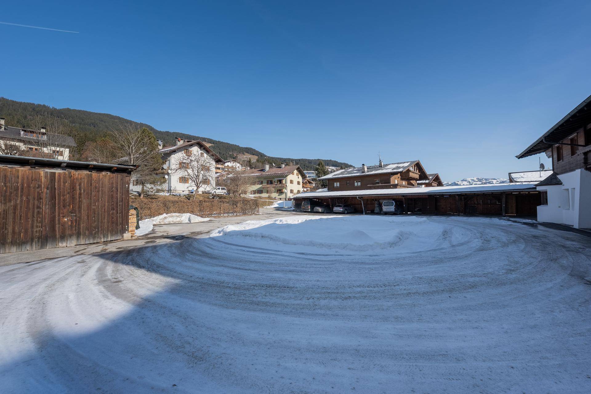 Blick auf den verschneiten Parkplatz vor traditionellen Gebäuden in einer ländlichen Umgebung mit Bergen im Hintergrund.