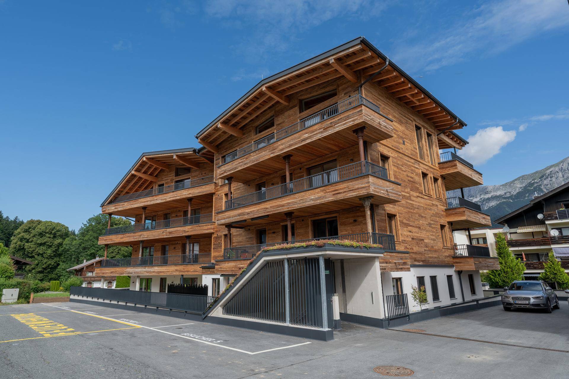 Neubau in Holzbauweise mit Balkonen und Tiefgaragenzufahrt, gelegen in einer bergigen Umgebung.
