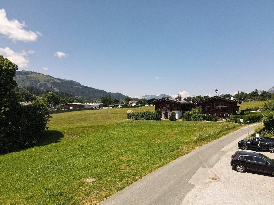 Blick über eine Straße und grüne Wiesen auf traditionelle Häuser und sanfte Hügel in der Ferne.