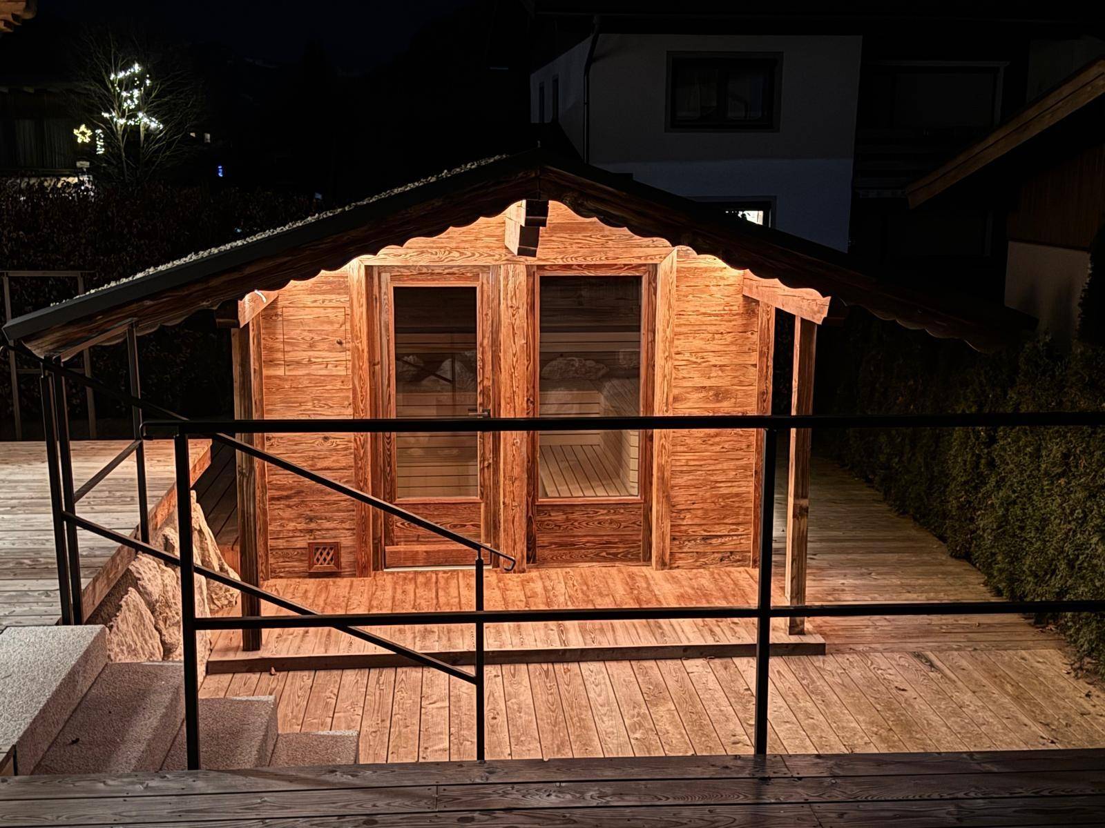 Externe Holzsaunakabine auf einer Holzterrasse, beleuchtet in der Nacht.