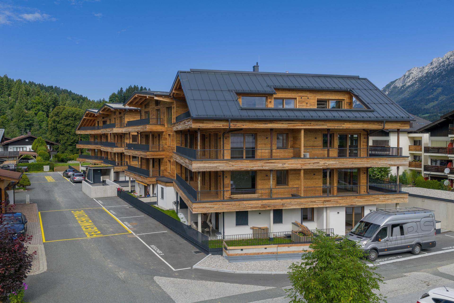 Zeitgenössisches Wohngebäude mit Holzverkleidung und Balkonen, umgeben von Parkplätzen und Bergkulisse.