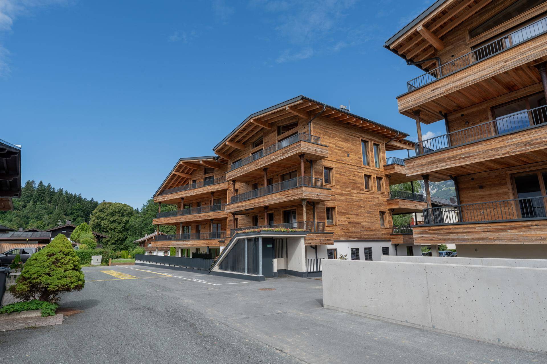 Mehrfamilienhaus mit Holzfassade und Balkonen, umgeben von Parkplätzen und grüner Landschaft.