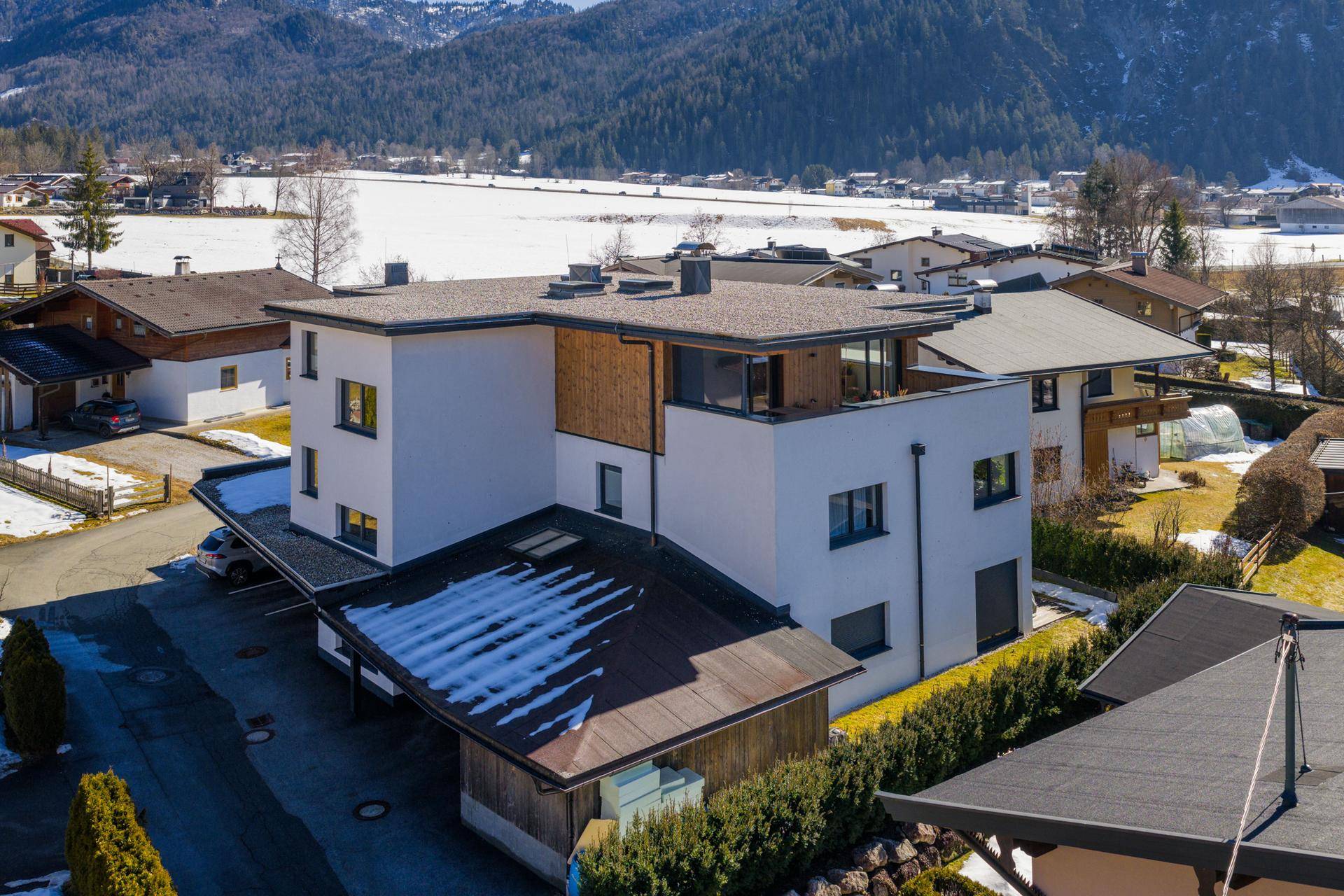 Moderne Wohnanlage mit Flachdach und Holzverkleidung, umgeben von verschneiten Feldern und Bergen.