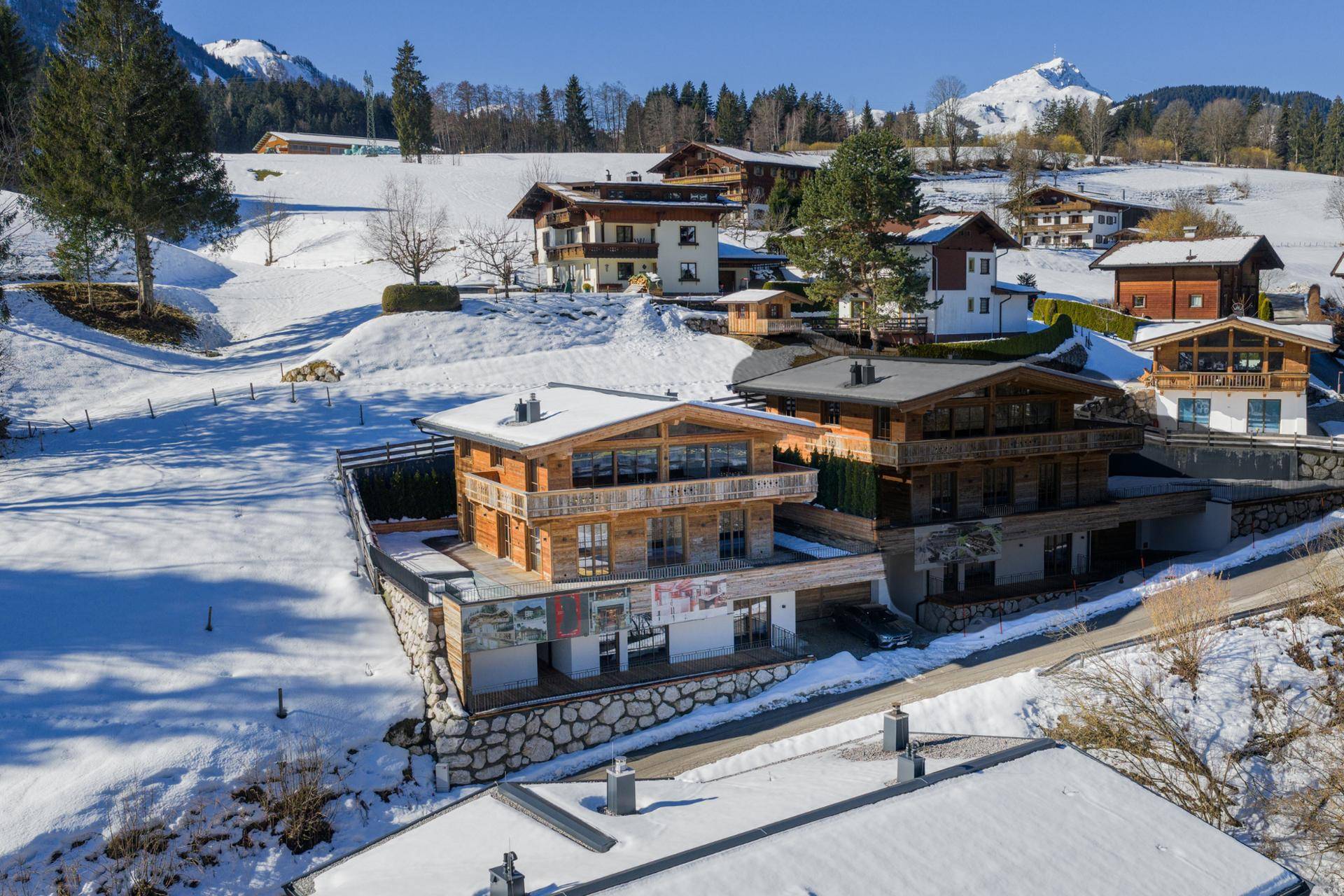 Panoramablick auf ein modernes Holzchalet in sonniger, verschneiter Umgebung mit Bergblick.