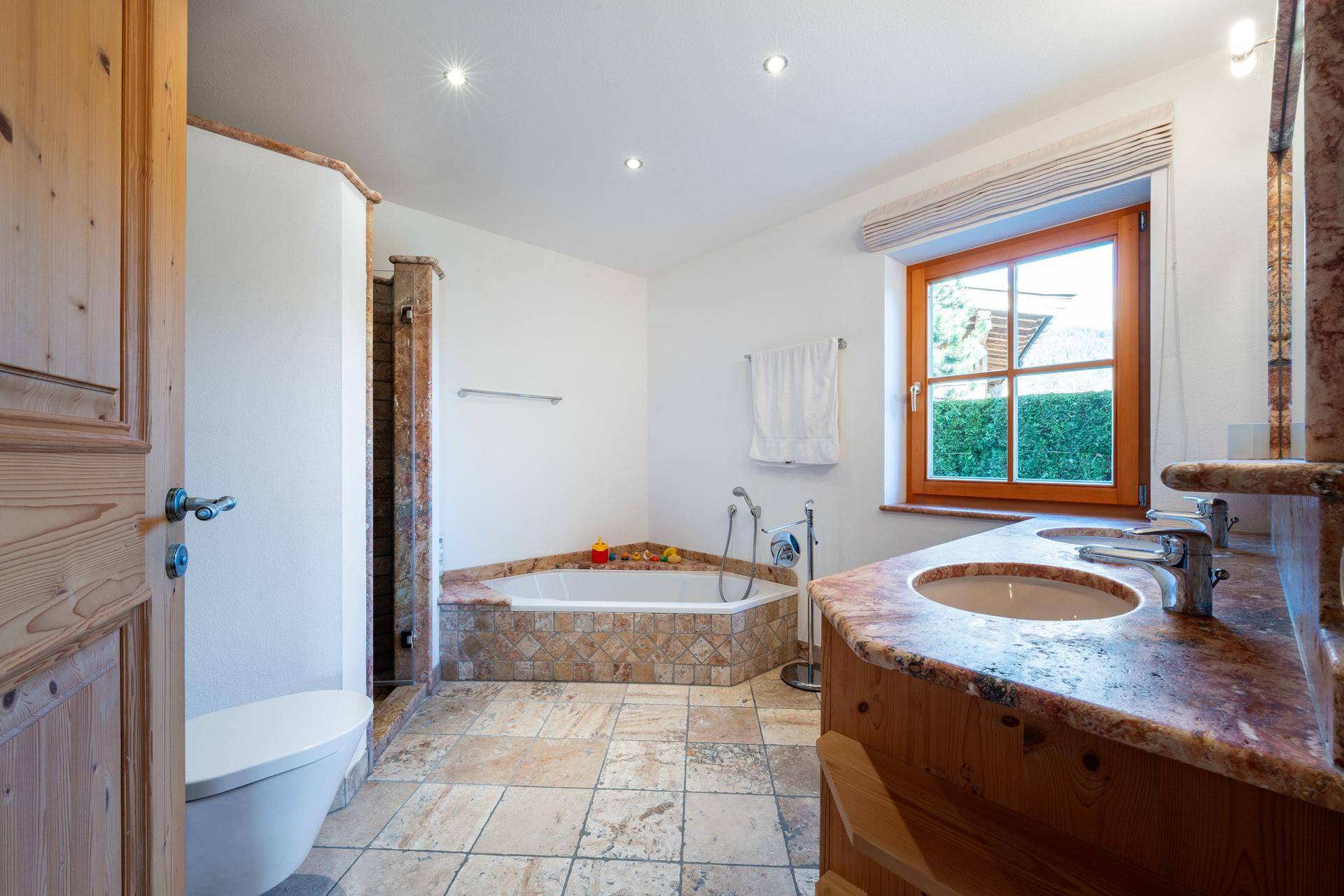 Badezimmer mit Eckbadewanne, Doppelwaschbecken und Fenster mit Blick ins Grüne.