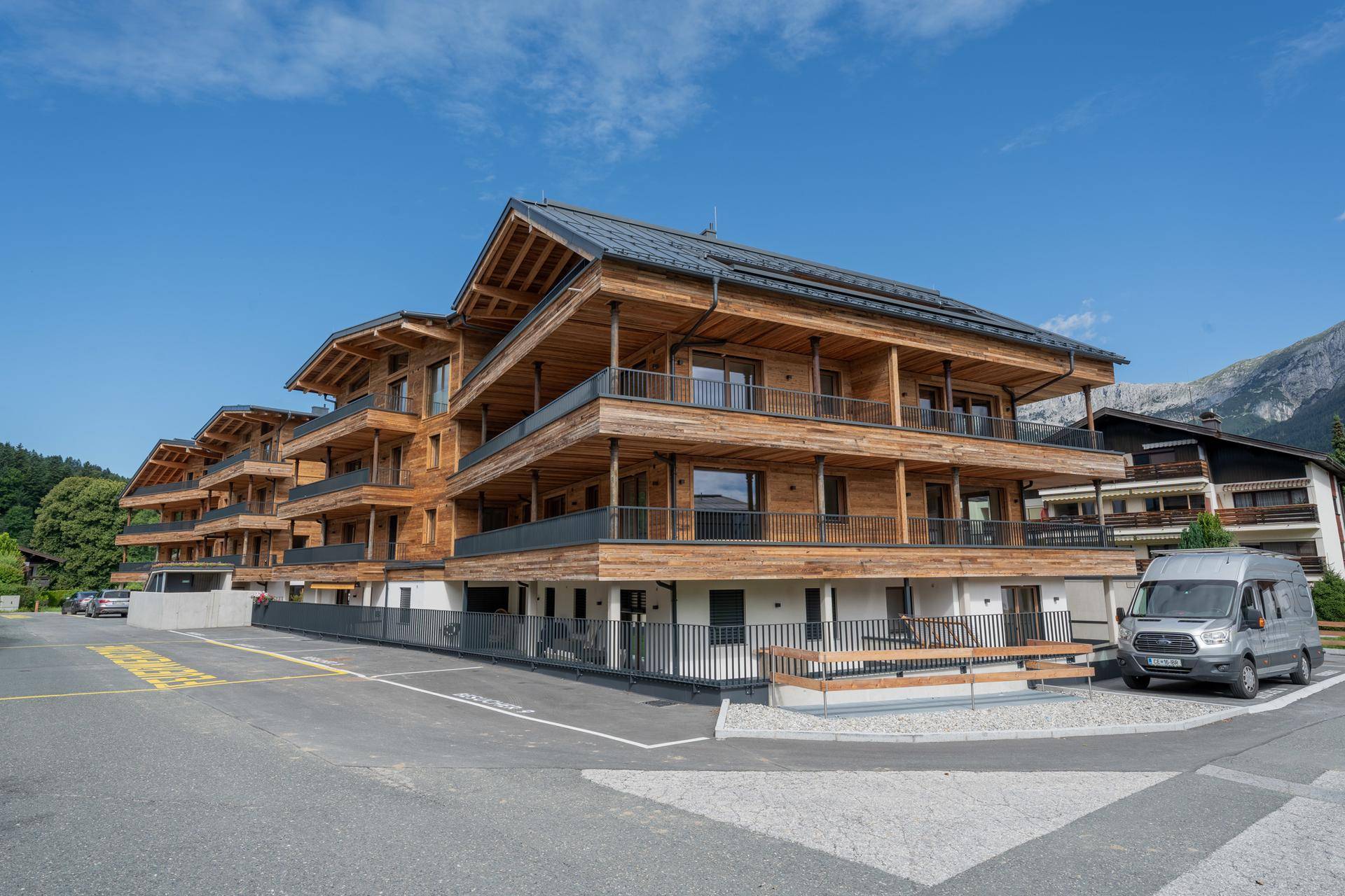 Attraktives Holzgebäude mit mehreren Etagen und Balkonen unter klarem blauem Himmel, mit Bergpanorama.