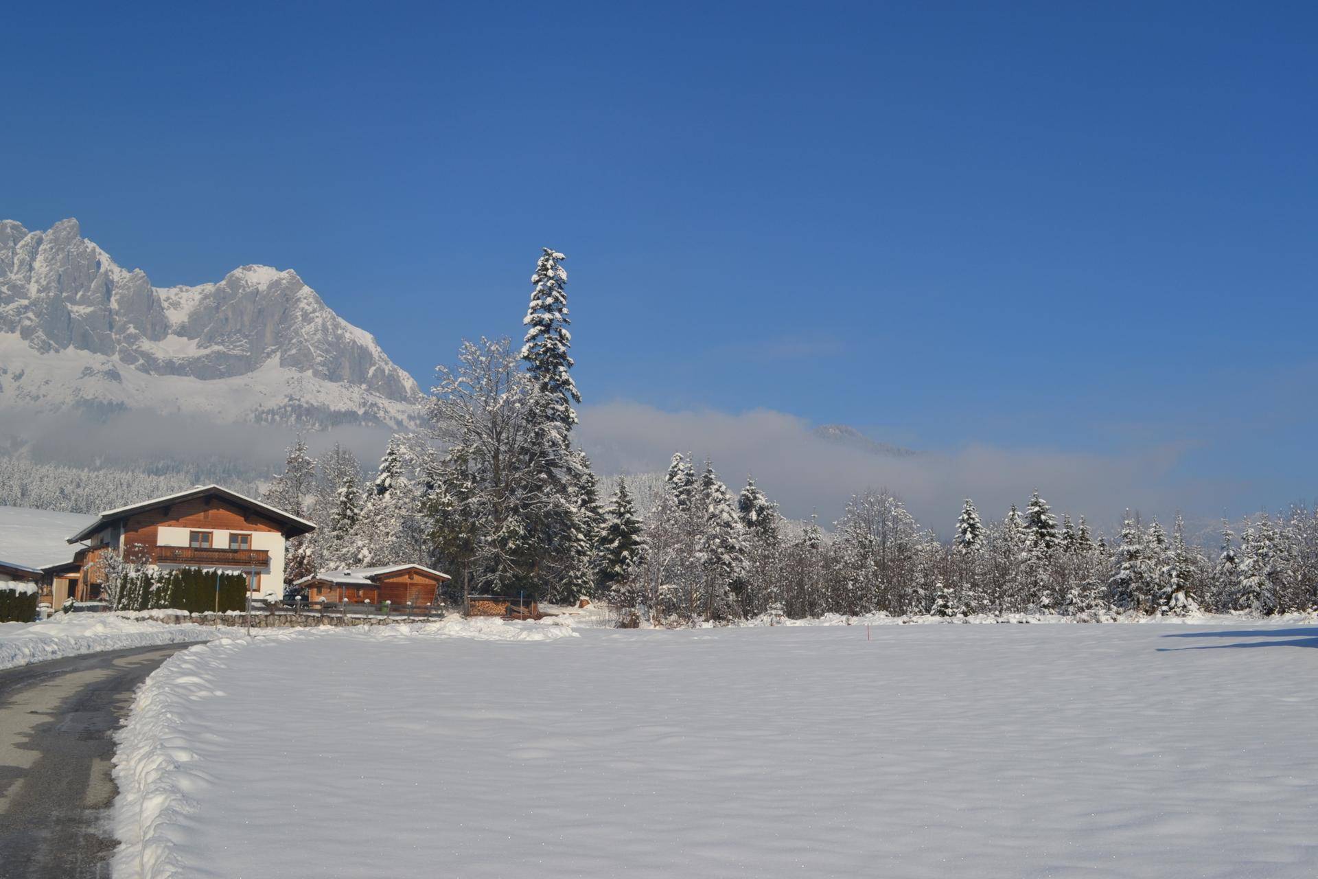 Winterlandschaft mit verschneiten Feldern, einem traditionellen Haus und schneebedeckten Bäumen vor Bergkulisse.