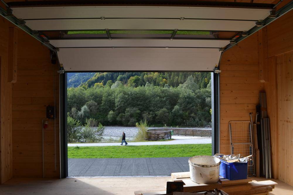 Blick aus einem geöffneten Rolltor auf eine grüne Landschaft mit Bäumen, einem Fluss und Bergen im Hintergrund.